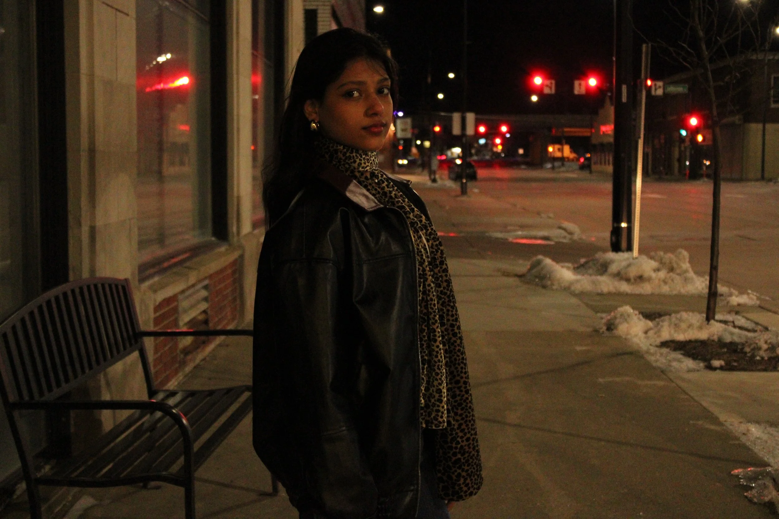 A woman standing on a sidewalk at night, wearing a black leather jacket and leopard print scarf, with red traffic lights and snow piles in the background.