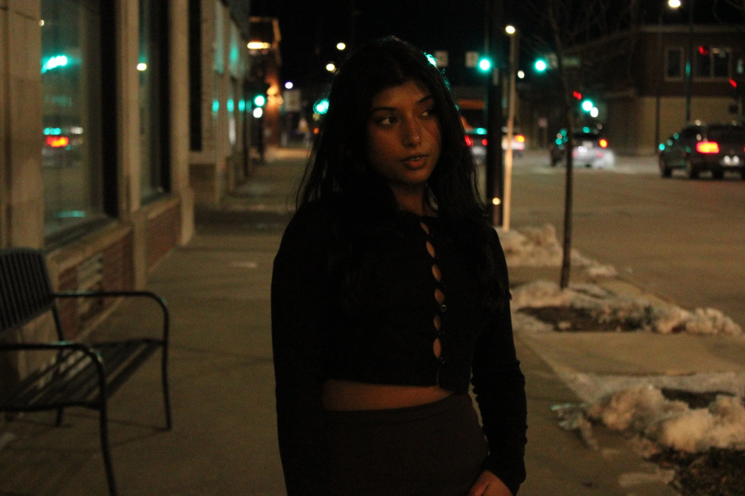 A woman with dark hair wearing a black jacket standing on a city sidewalk at night, with snow on the ground and illuminated streetlights and traffic lights in the background.