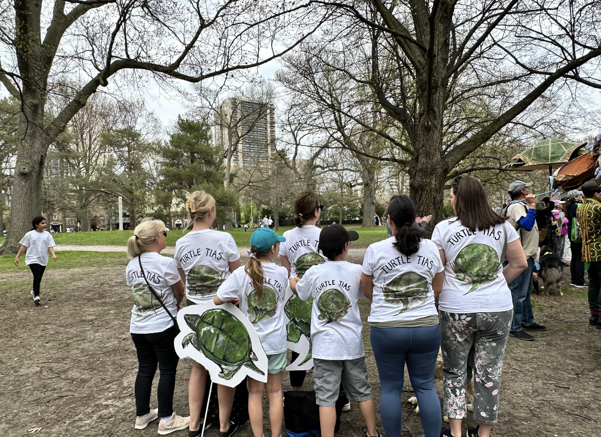 Group of people gathered outdoors in a park, wearing T-shirts with a turtle illustration and the words "TURTLE TIAS.
