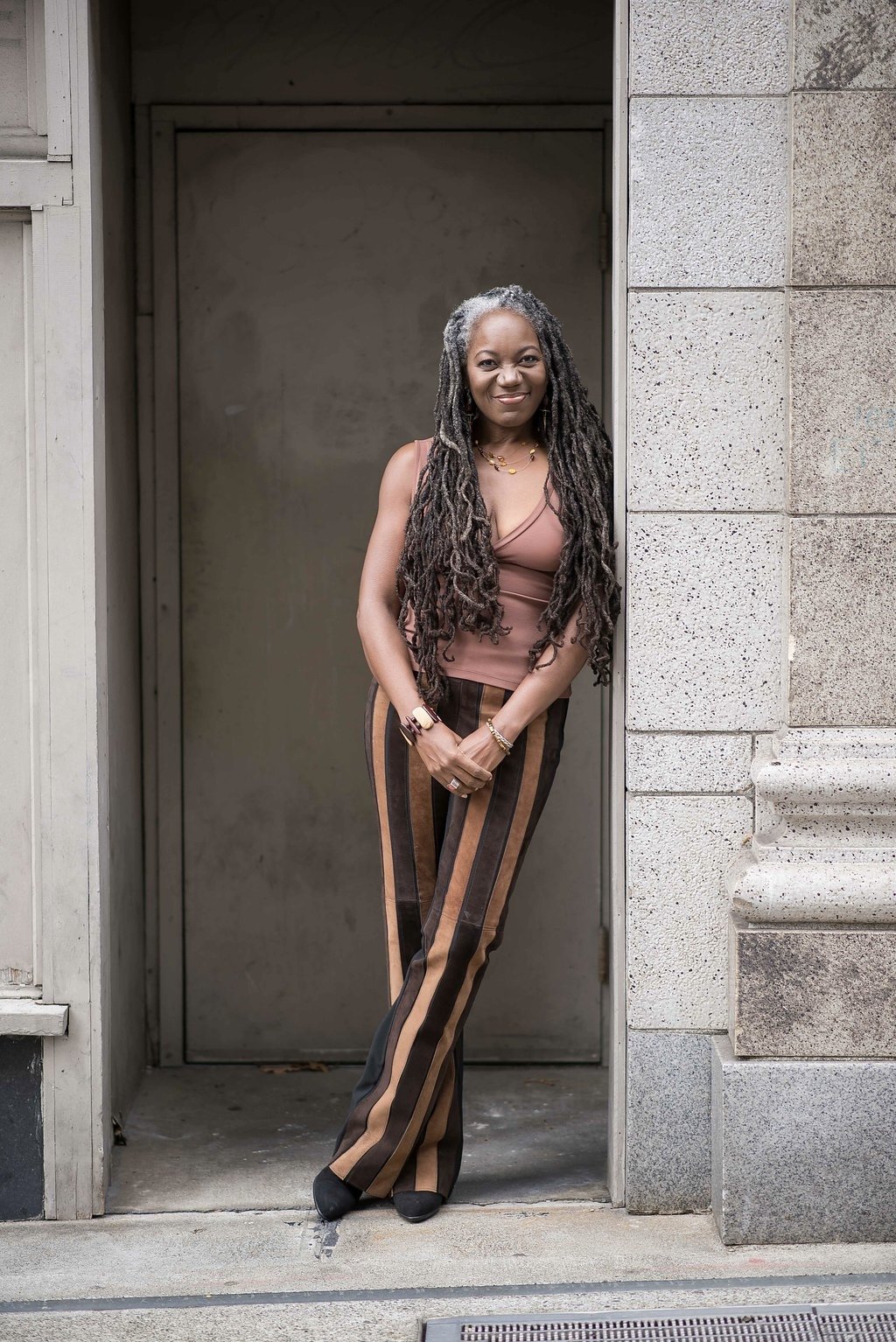 A woman with long dreadlocks stands in a doorway, wearing a sleeveless brown top and striped pants, with jewelry on her hands and neck.