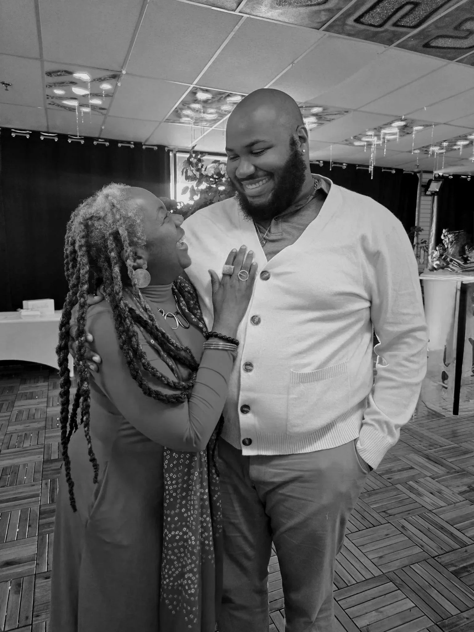 A joyful woman with gray hair and earrings touches the chest of a smiling, bald man with a beard, dressed in a light cardigan and pants, in an indoor setting with a decorated ceiling, a window with curtains, and a patterned wooden floor.