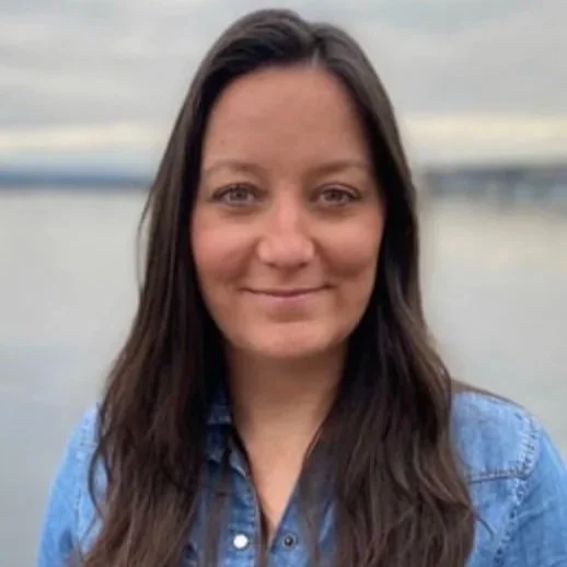 A woman with long brown hair, wearing a blue denim shirt, standing outdoors near a body of water under an overcast sky.
