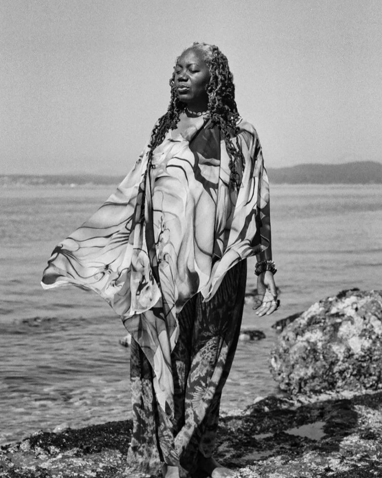 A woman with curly hair standing on rocks by the water, wearing a patterned shawl and loose pants, with her eyes closed.