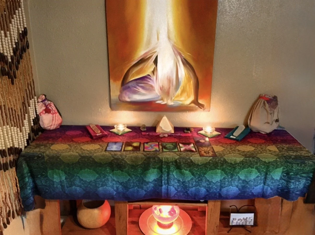 A table decorated with a colorful cloth, tarot cards, candles, and crystals, set against a wall with an artistic painting of a person, and adorned with items such as bags and books.
