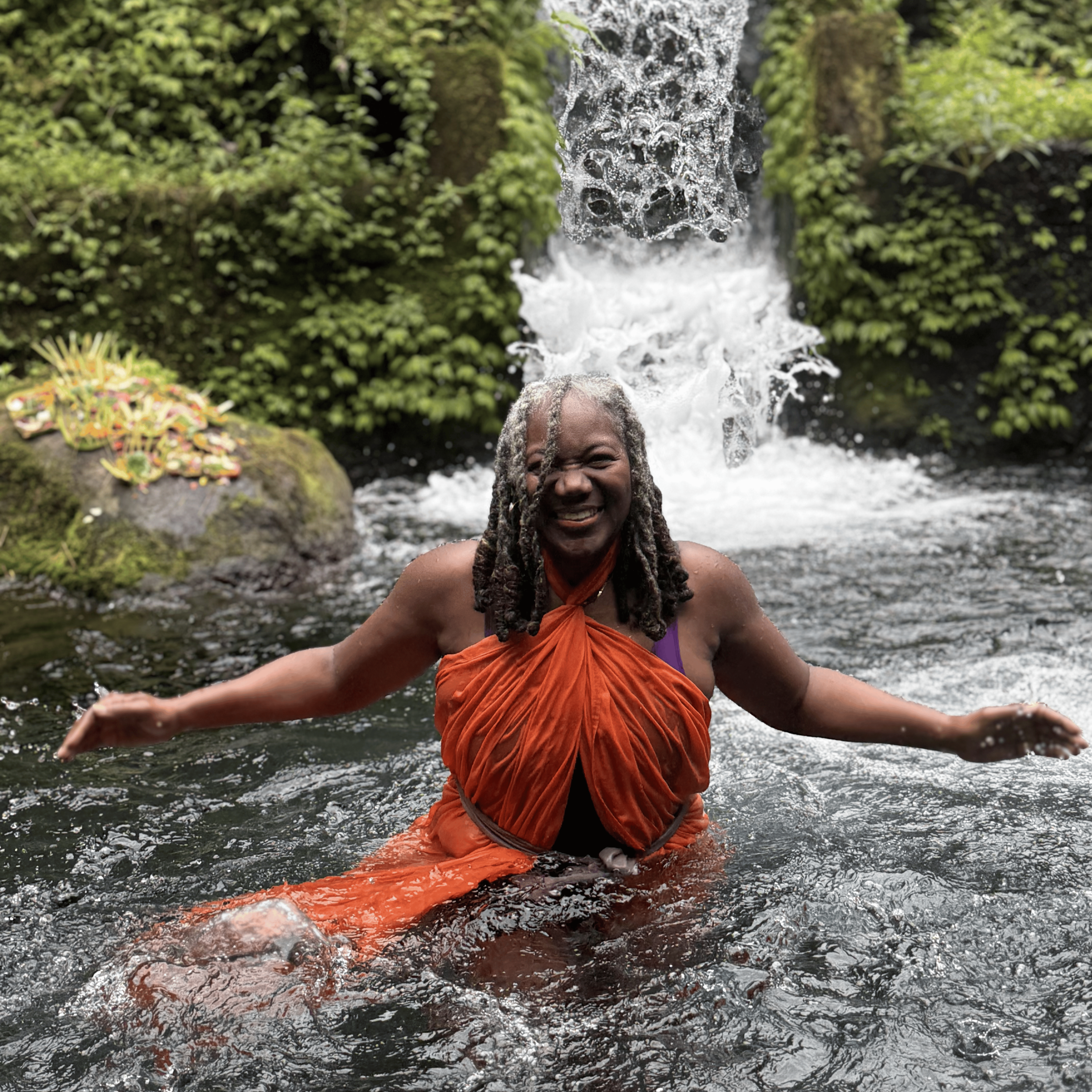 Woman with shoulder-length dreadlocks smiling and standing in a pond near a small waterfall in a lush, green forest.