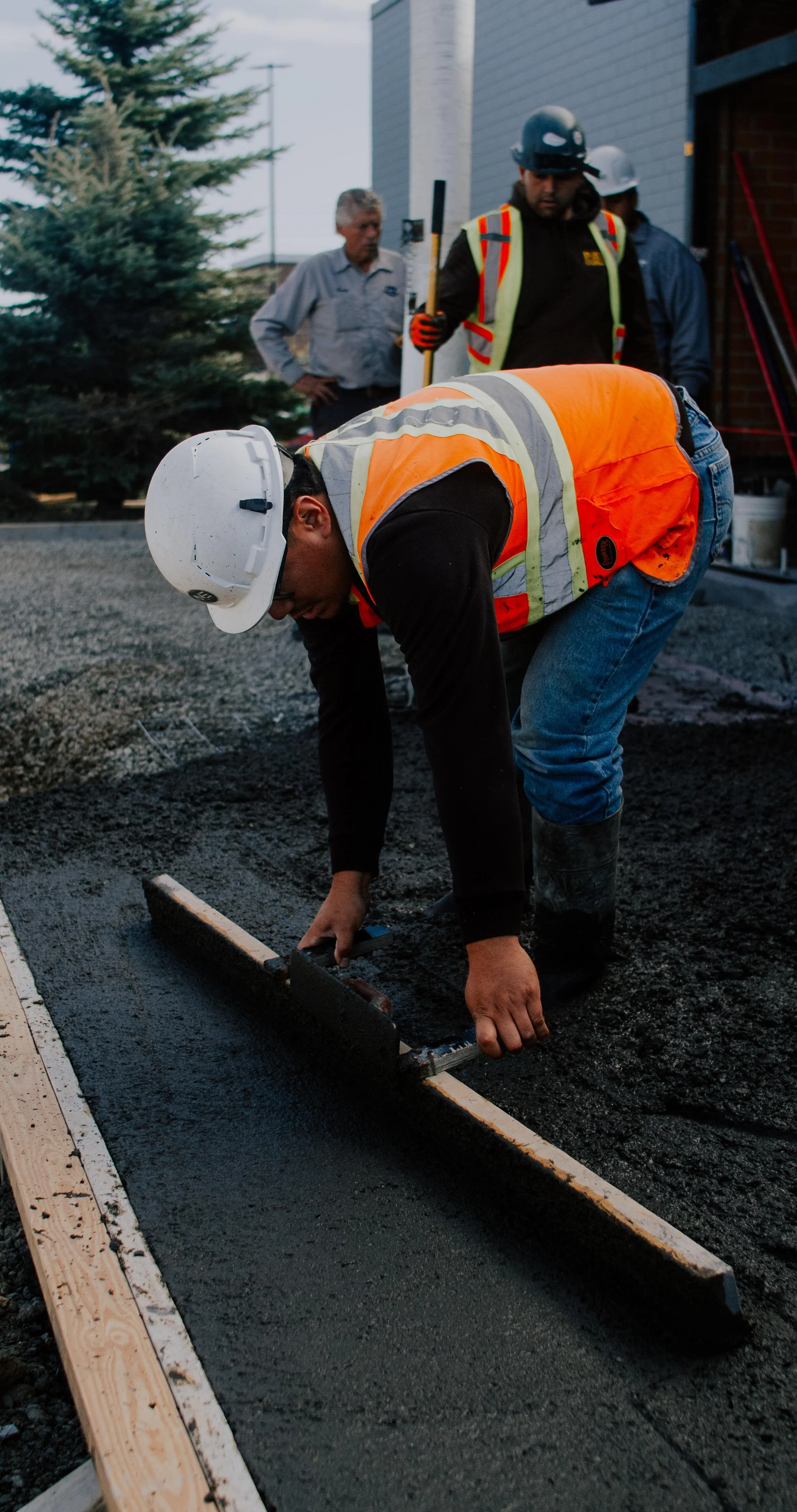 Construction worker in an orange safety vest and white helmet smoothing wet concrete with a tool, while two other workers in helmets and safety gear stand in the background near a building.