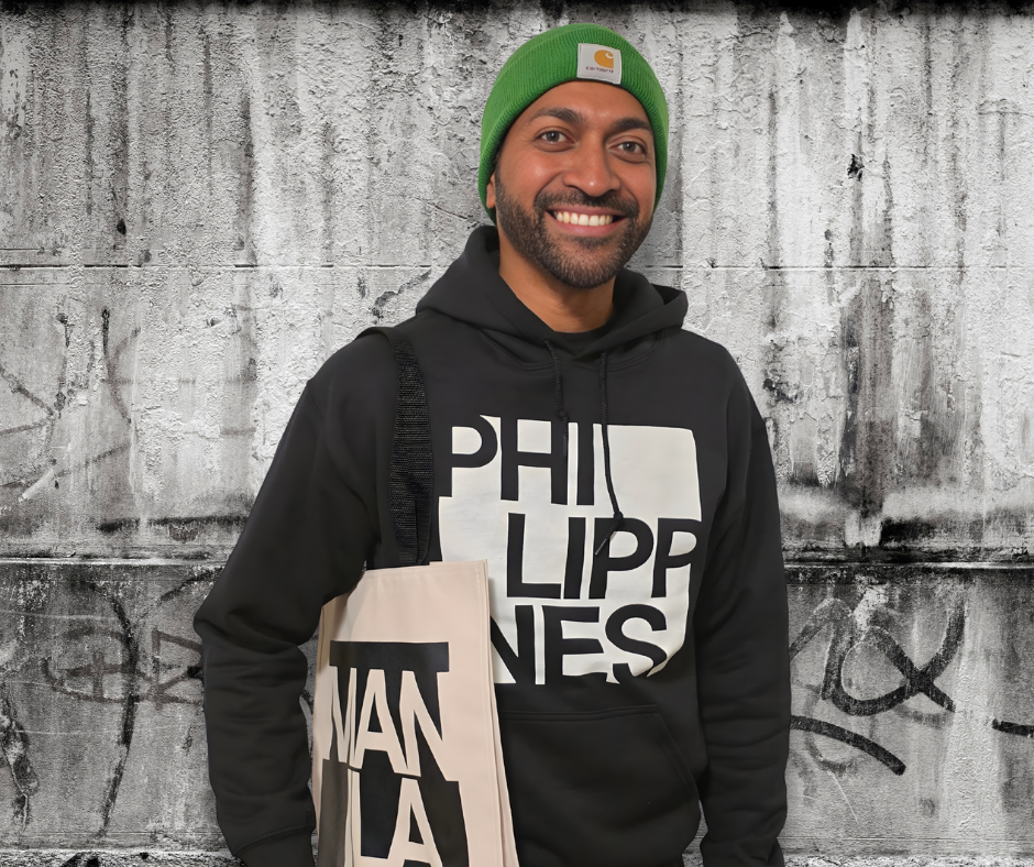 A man smiling, wearing a green beanie and a black hoodie with the text 'PHILIPPINES' on it, standing in front of a textured concrete wall with graffiti, holding a cream tote bag.