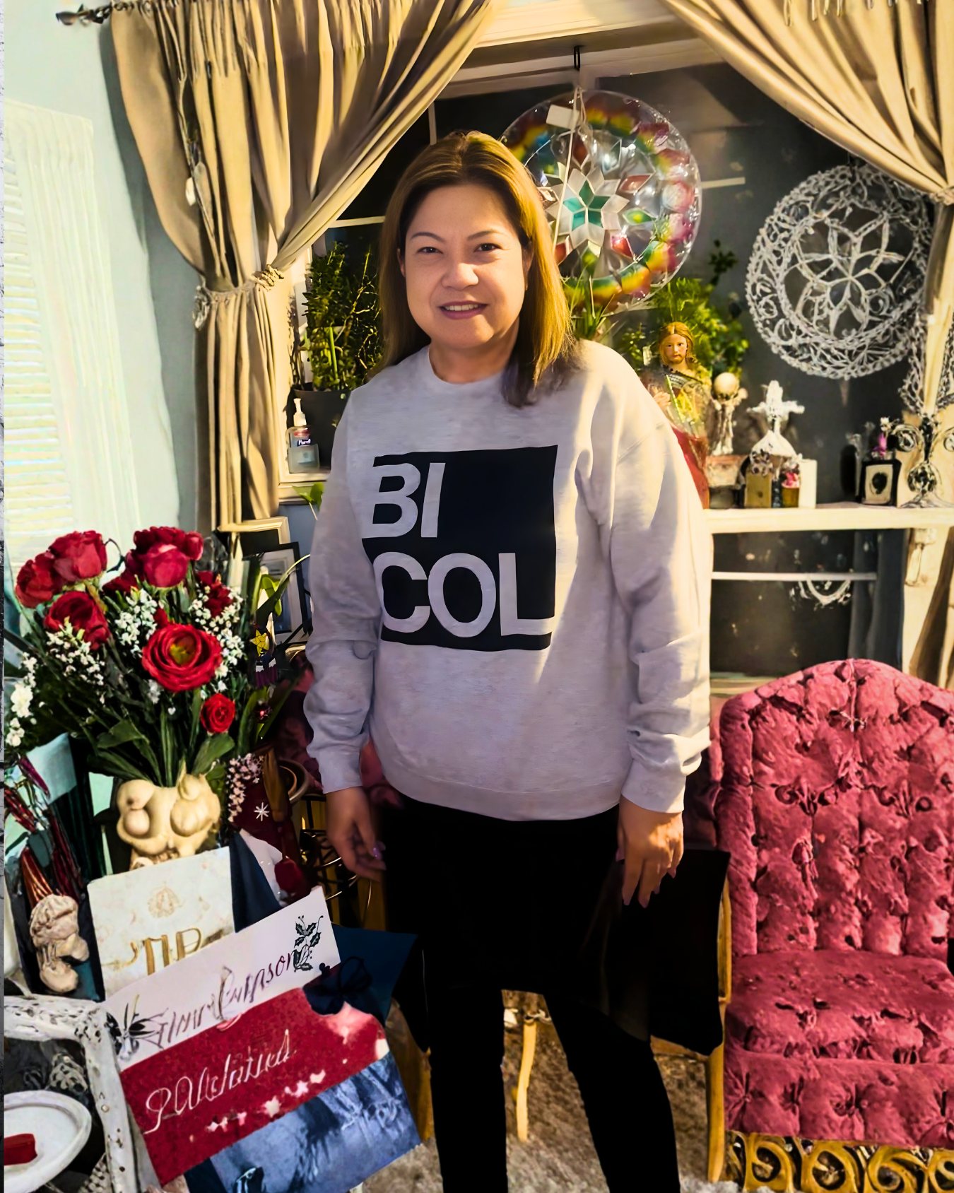 A woman standing in a room decorated with various ornaments, including a bouquet of red flowers, a pink velvet chair, and decorative items on a white shelf, with gold and beige curtains in the background.