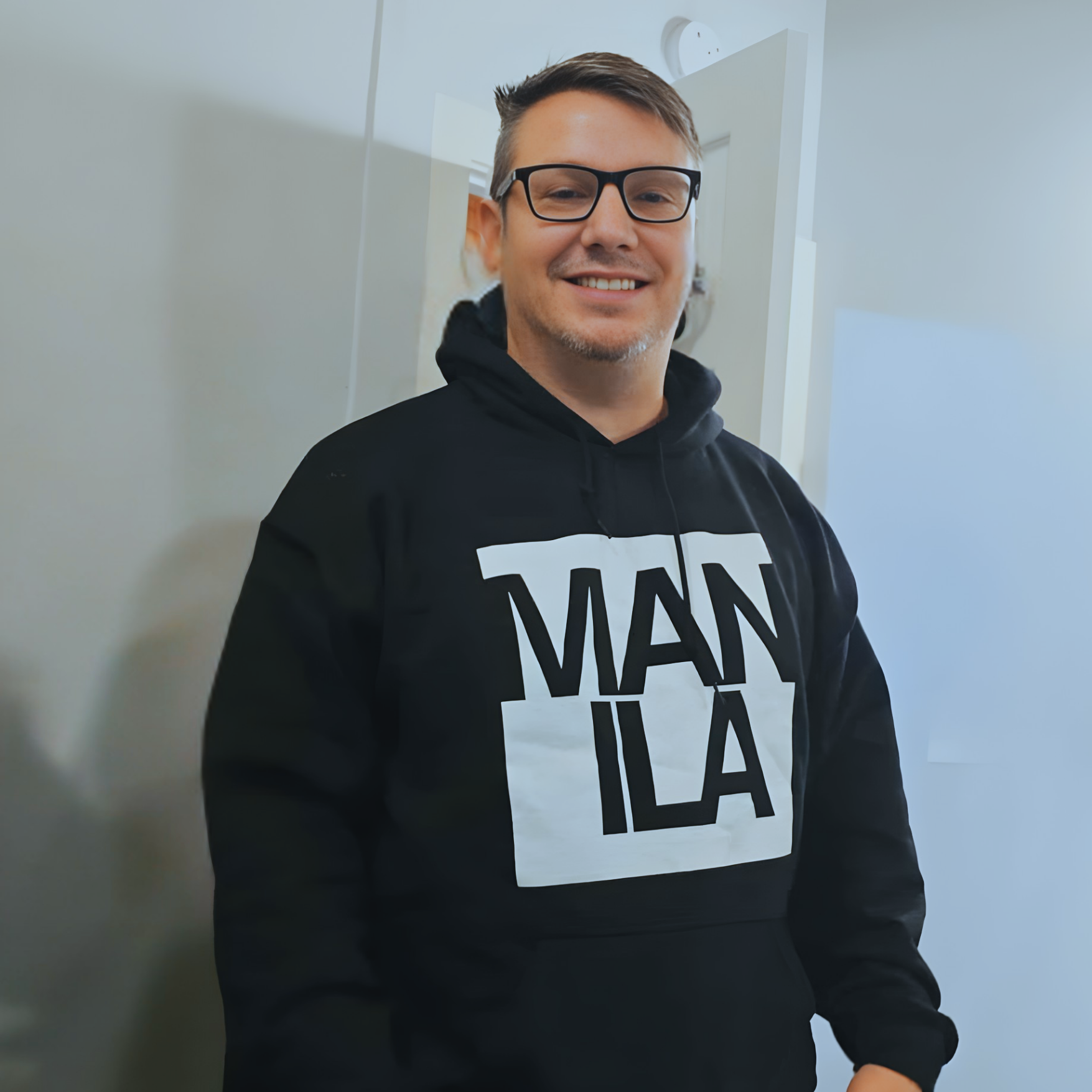 A man with glasses and short brown hair, smiling, wearing a black hoodie with the words 'MAN I LA' printed on it, standing indoors against a light-colored wall.