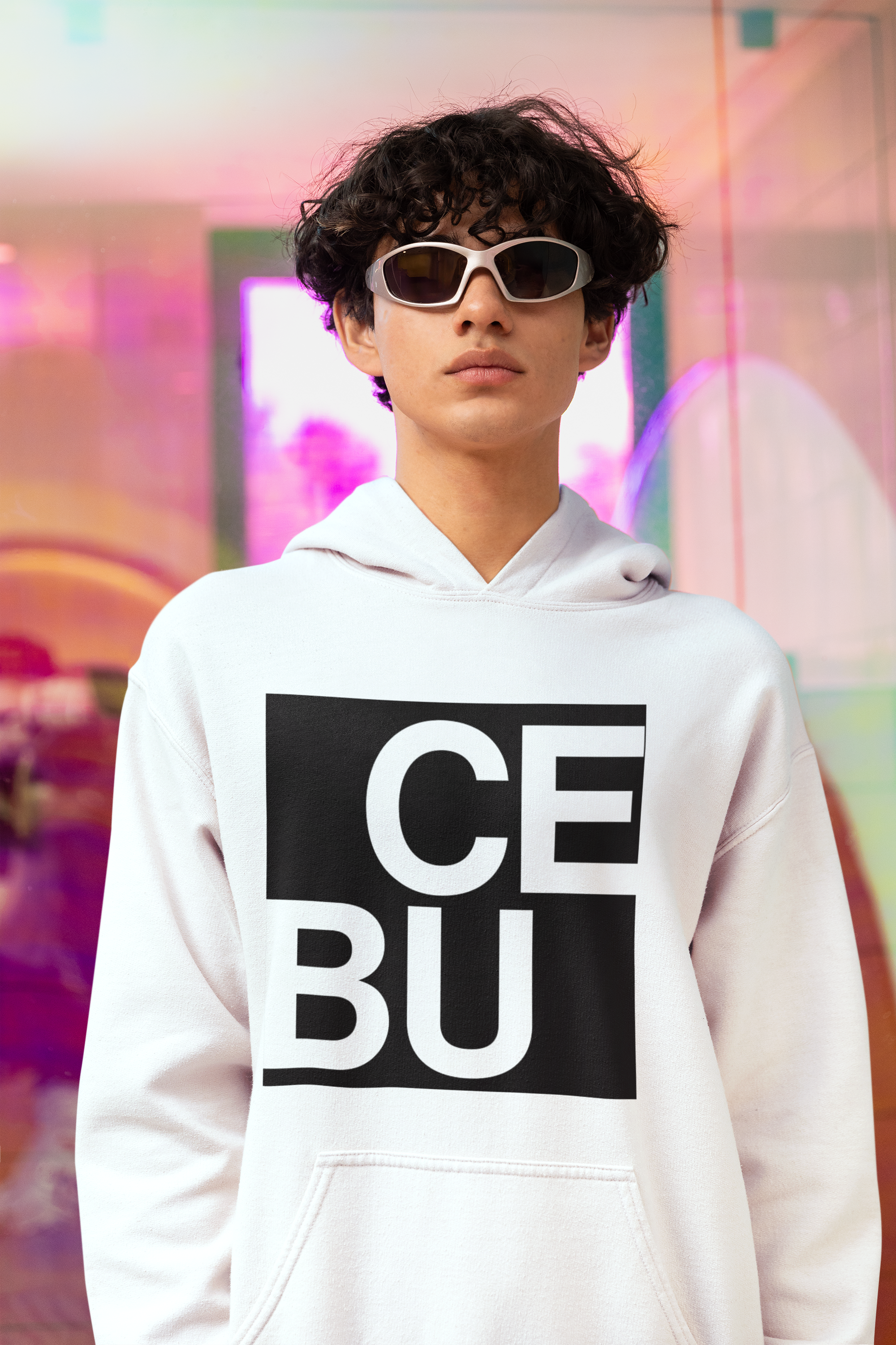 A young man with dark curly hair wearing sunglasses and a white hoodie with the word "CEBU" printed in black on the front, standing in front of a colorful, reflective background.