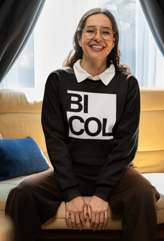 A young woman with glasses and long brown hair sitting on a beige sofa in front of window with curtains, smiling at the camera. She wears a black sweatshirt with the word 'BICOL' printed on it and a white collared shirt underneath.
