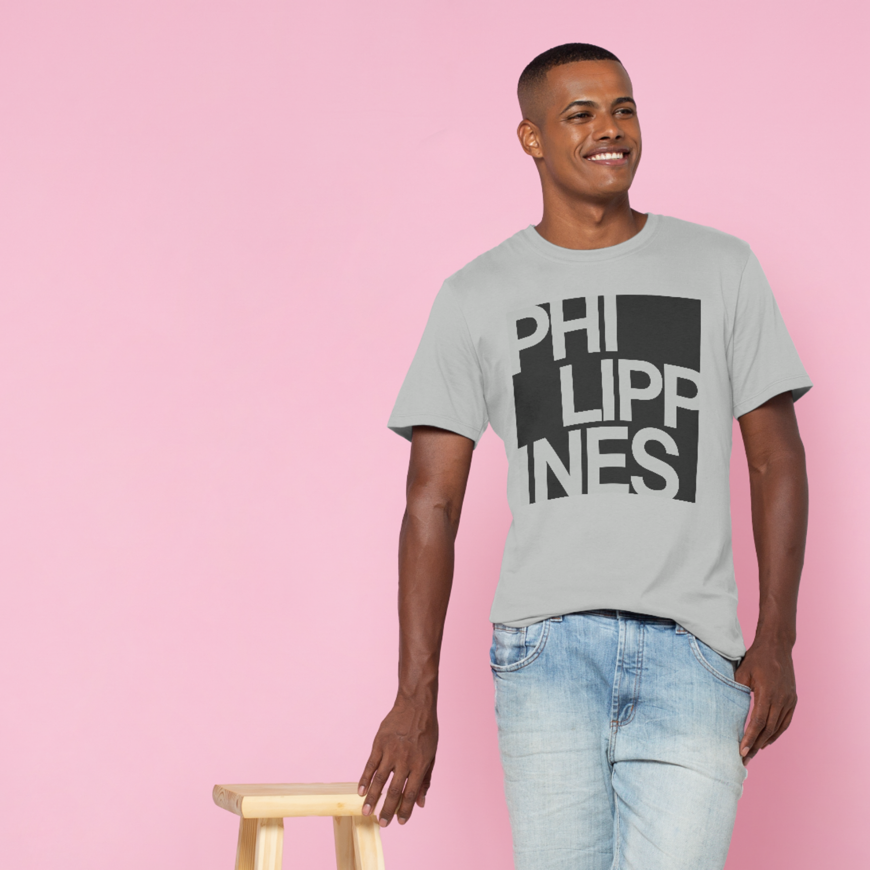 Young man with short hair smiling, wearing a light gray T-shirt with a black and gray geometric graphic and text, and light blue jeans, standing against a pink background, holding a wooden stool with his right hand.