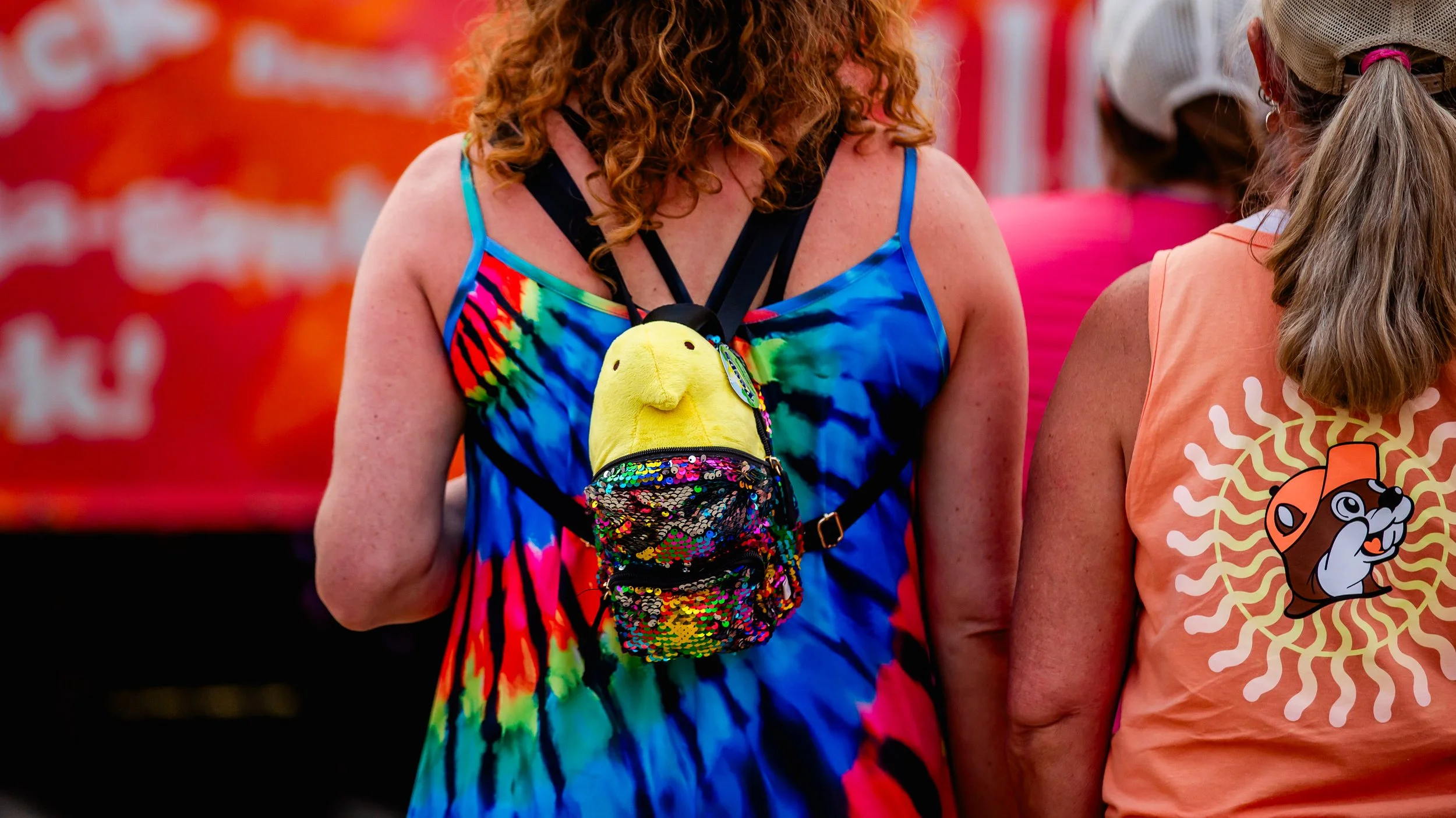 People at an outdoor event, one with a tie-dye dress and a colorful backpack with a smiley face, and another person with a sleeveless top featuring a cartoon dog design.