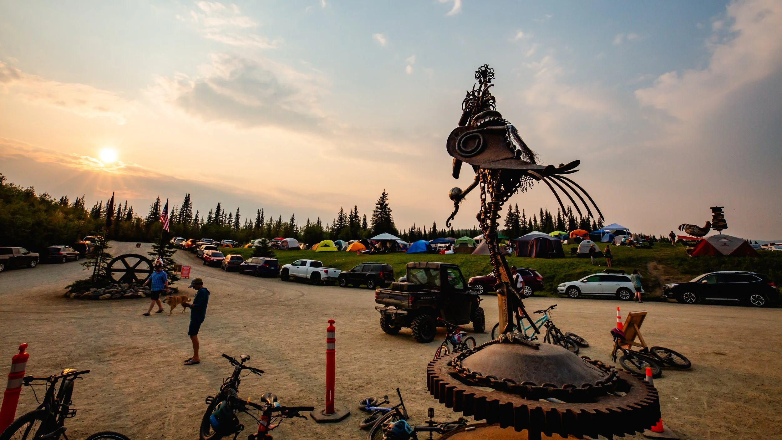 An outdoor scene during sunset with a large metal sculpture of an insect, surrounded by cars, tents, and people. Some bicycles and a utility vehicle are also visible. The background features a forested area and a cloudy sky.