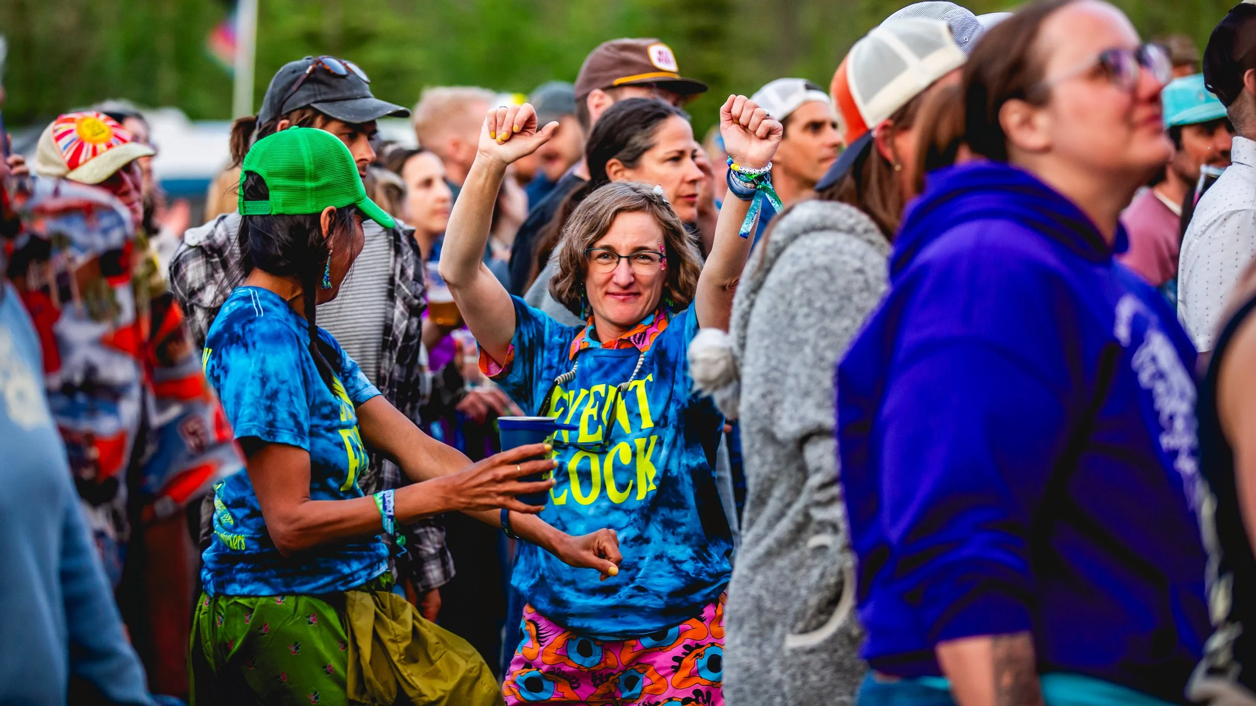 Crowd of people at outdoor event, woman in colorful outfit with raised arms, smiling, wearing glasses, surrounded by others in casual attire and hats.