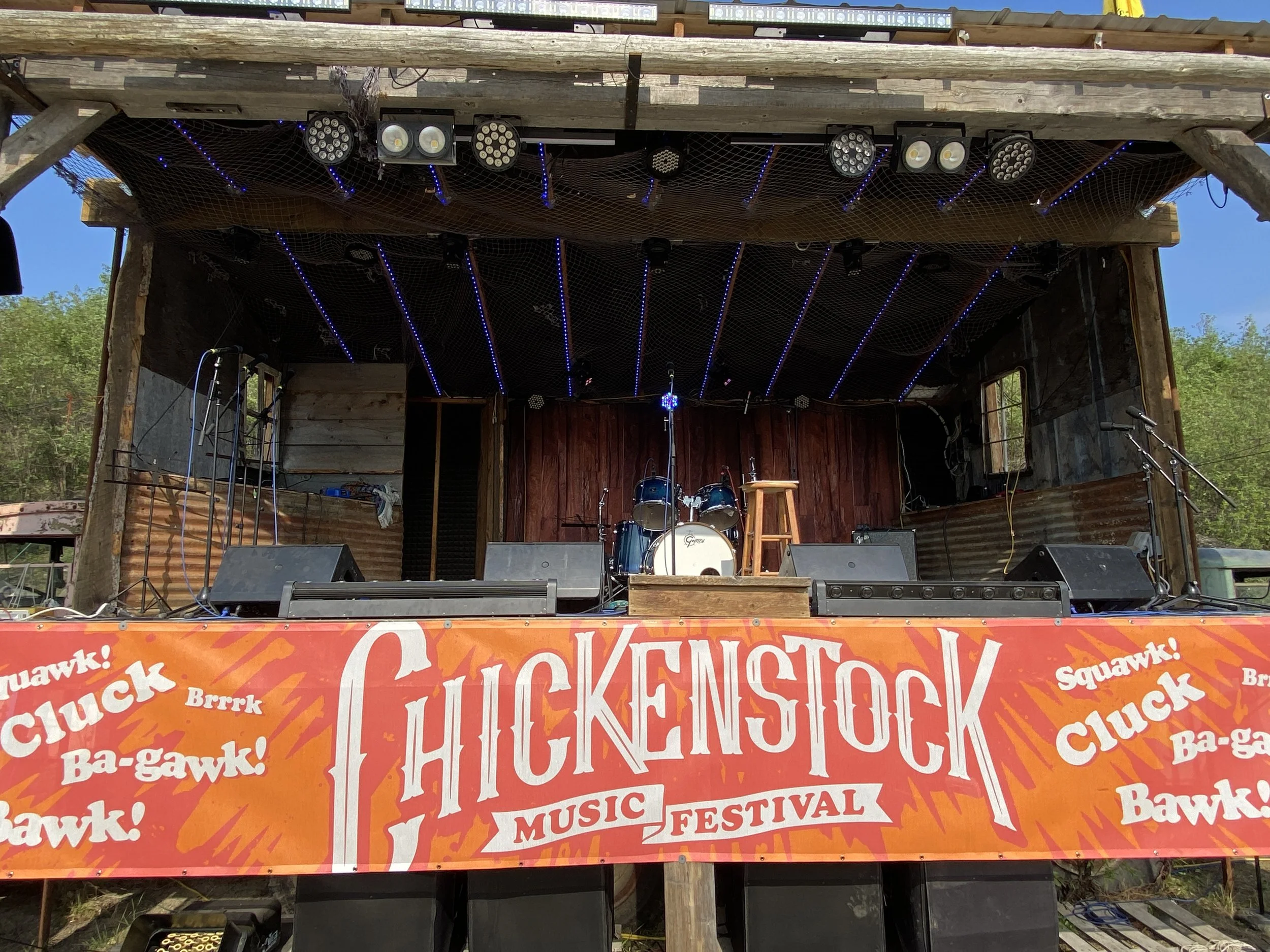 Empty stage at the Chickfest Music Festival with drums, a stool, and microphones, under a rustic wooden shelter, with a bright orange banner featuring event branding.