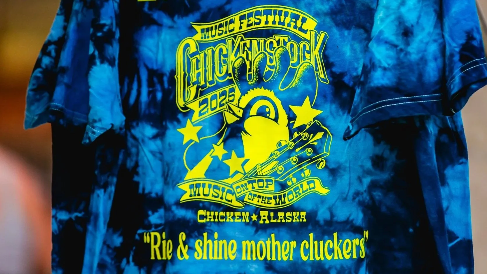 Blue tie-dye T-shirt with a colorful graphic promoting Chuckfest 2025 music festival in Chicken, Alaska, featuring a cartoon duck, stars, a guitar, and the slogan 'Rie & shine mother cluckers'.