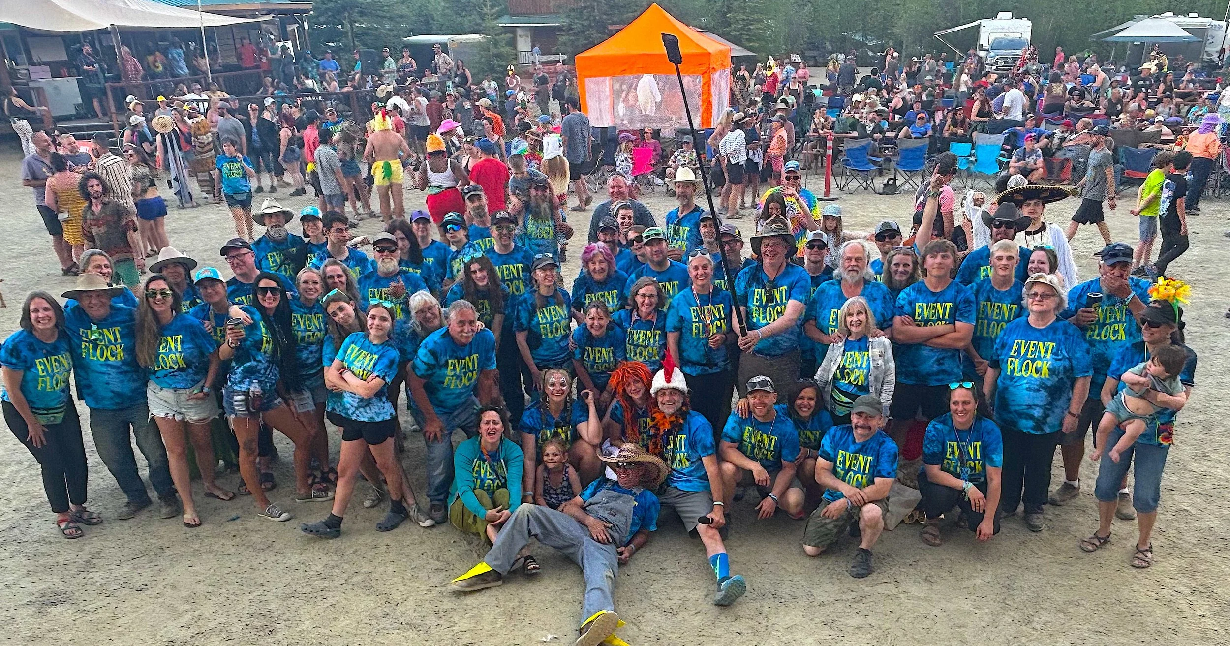 Group of people at an outdoor event, many wearing matching blue shirts with 'Event Flock' printed on them, some wearing hats and accessories, posing for a group photo in front of a crowd at a festival or fair.
