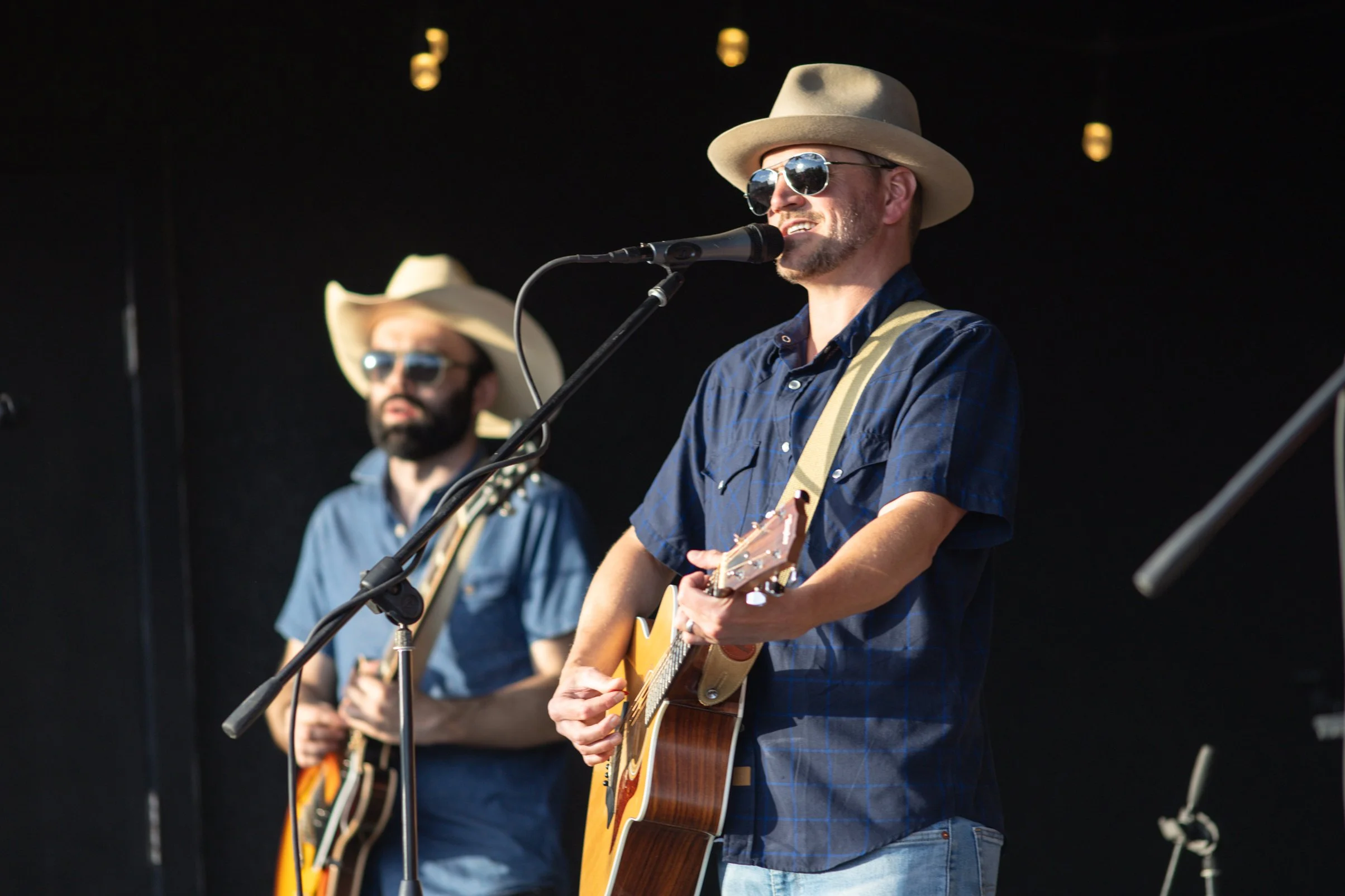 Two men performing on stage, both wearing cowboy hats and sunglasses. One is singing into a microphone while playing an acoustic guitar, and the other is holding a different guitar in the background.