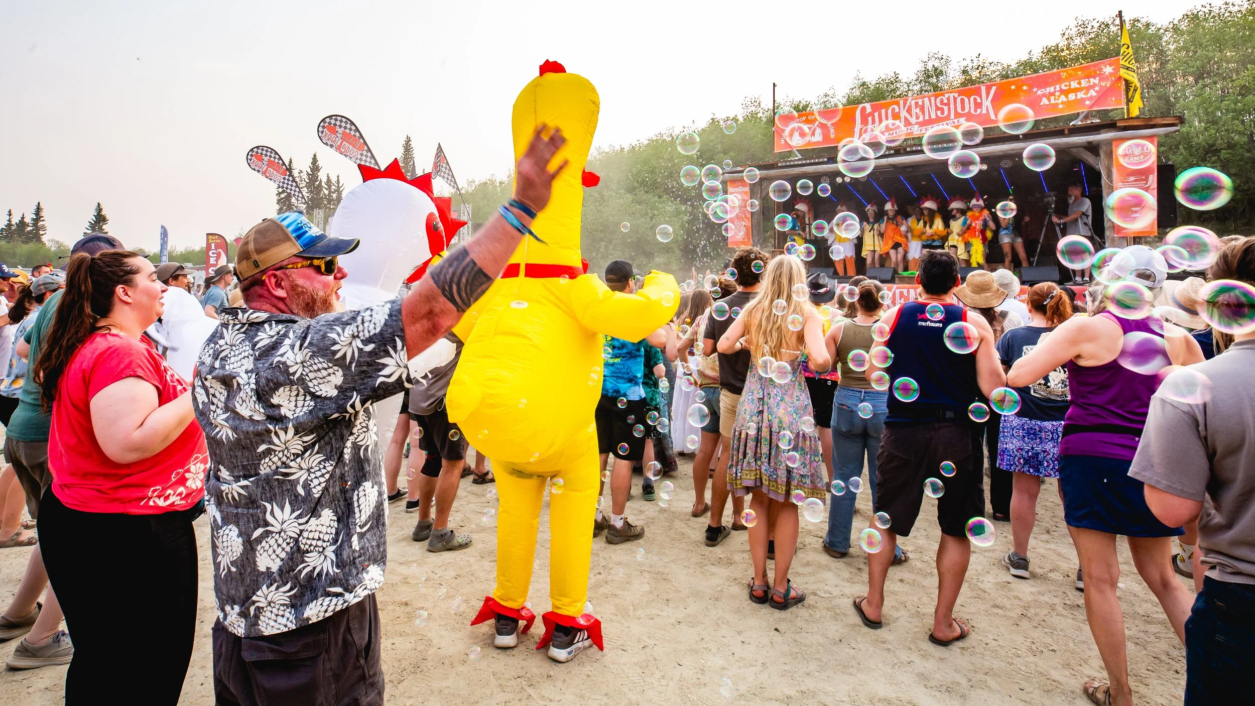 Crowd of people enjoying an outdoor concert with bubbles, a stage with performers, and a person in a yellow chicken costume at a festival.