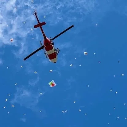 Aerial view of a helicopter flying in a clear blue sky with scattered small papers or flags.
