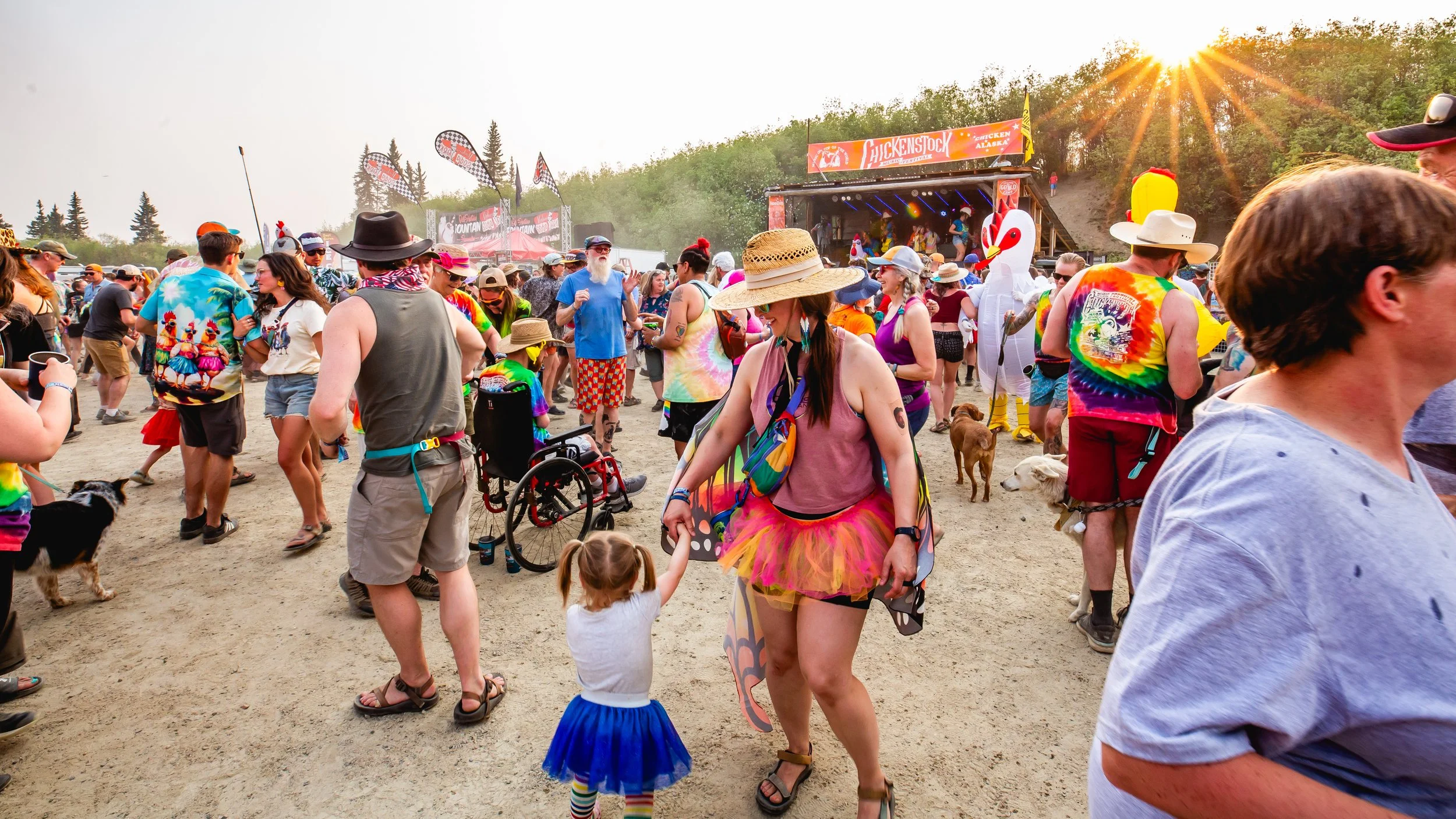 Crowd at an outdoor festival or concert during daytime with many people, some in colorful and tie-dye clothing, some with costumes, and a stage in the background with a large inflatable chicken and a banner that says "Hickensock." The sun is shining brightly, and people are enjoying the event.