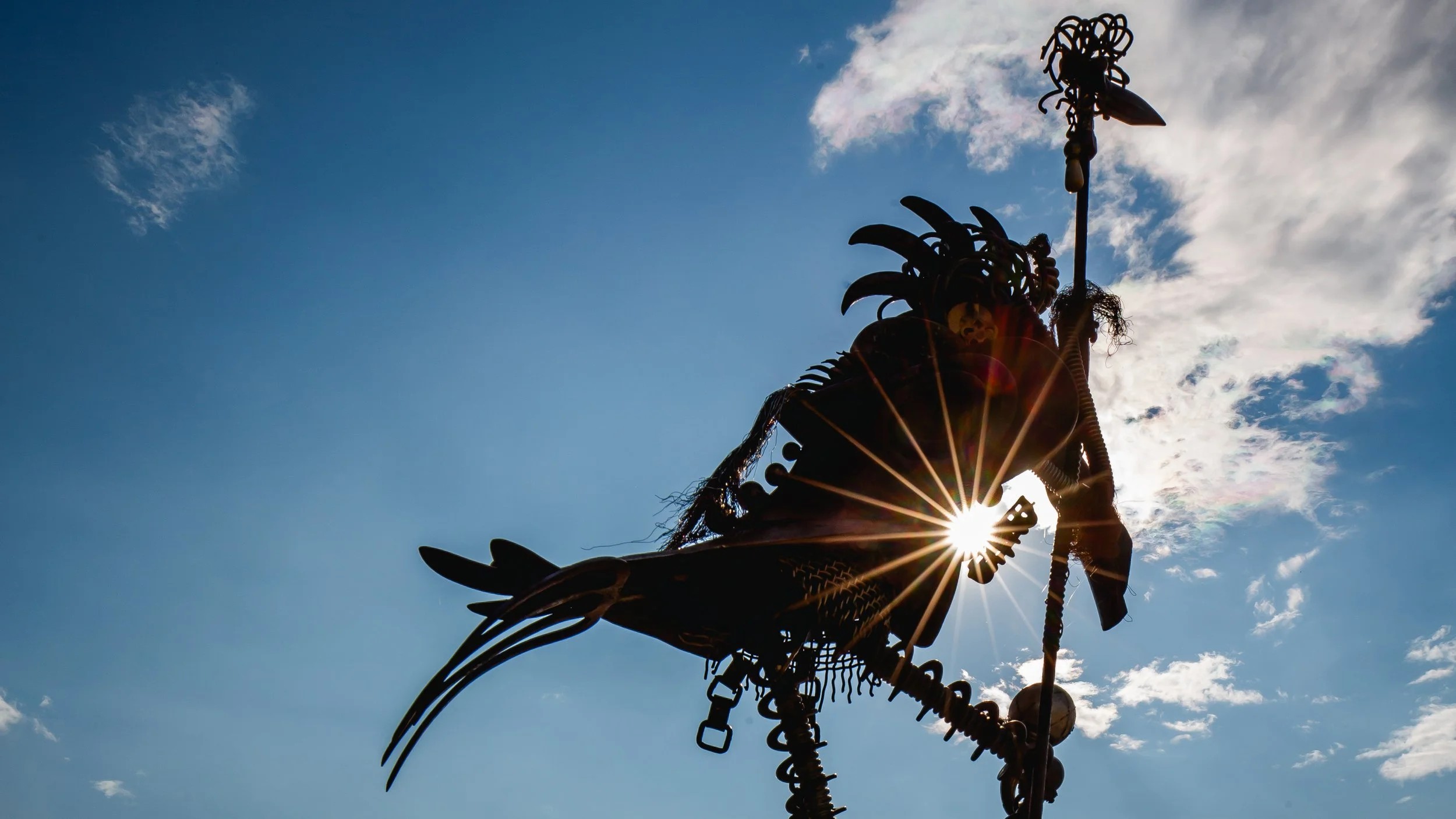 Silhouette of a metallic sculpture of a warrior holding a spear, with the sun shining through the sculpture against a blue sky with scattered clouds.