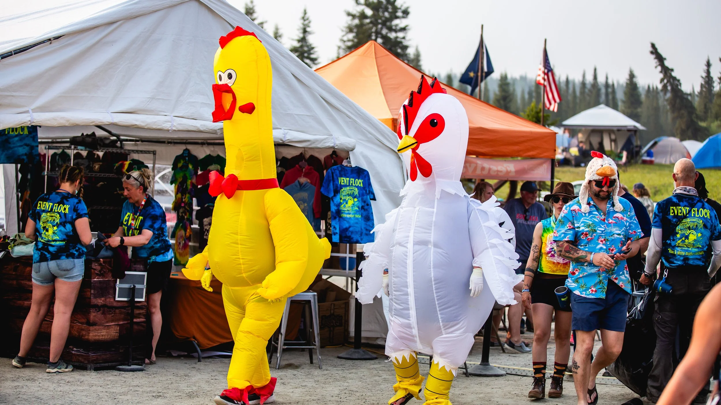 People in chicken costumes, booths, and festival tents at an outdoor event with trees and flags in the background.