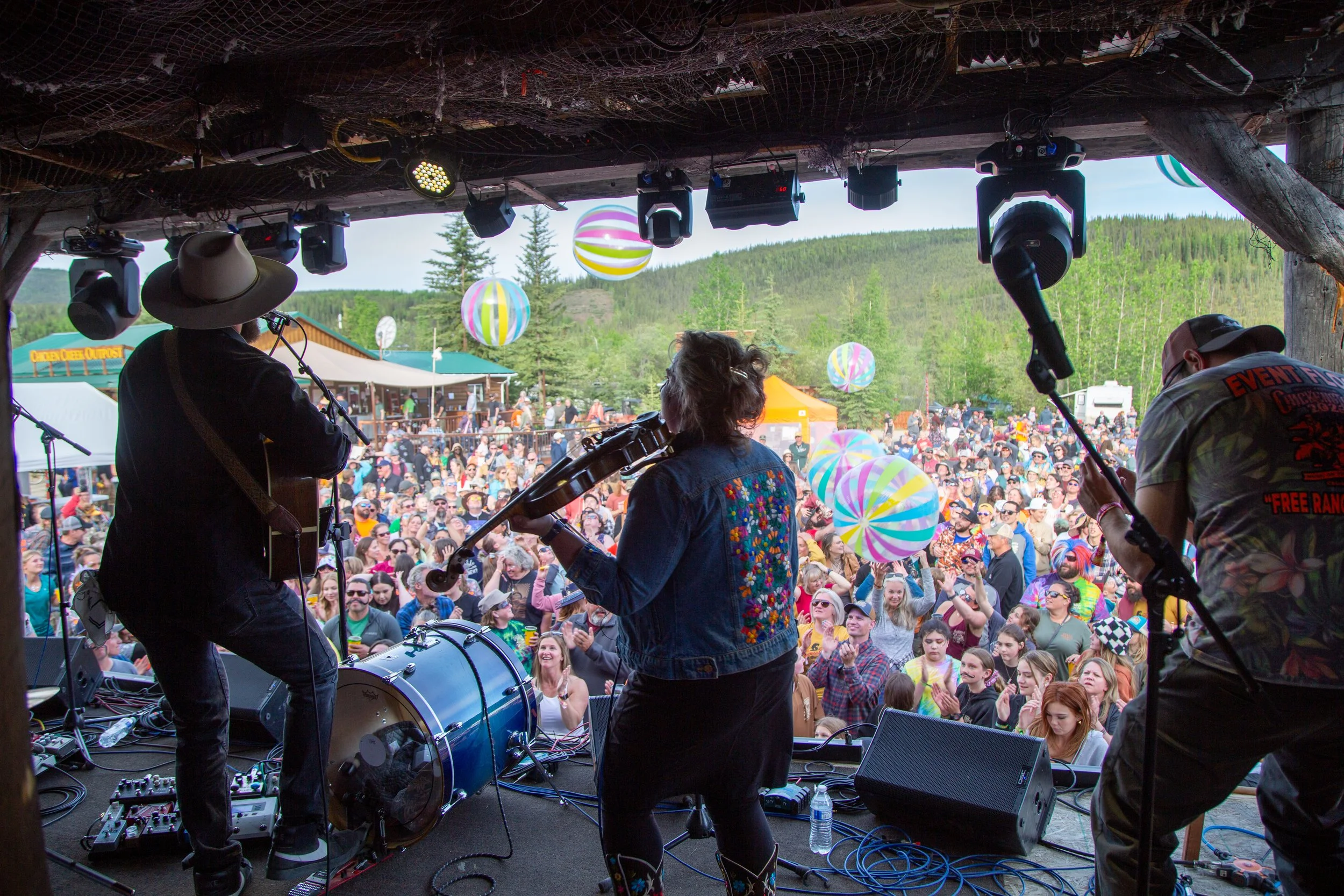 Musicians performing on stage at an outdoor music festival with a crowd of festival-goers holding colorful balloons in a mountainous area.