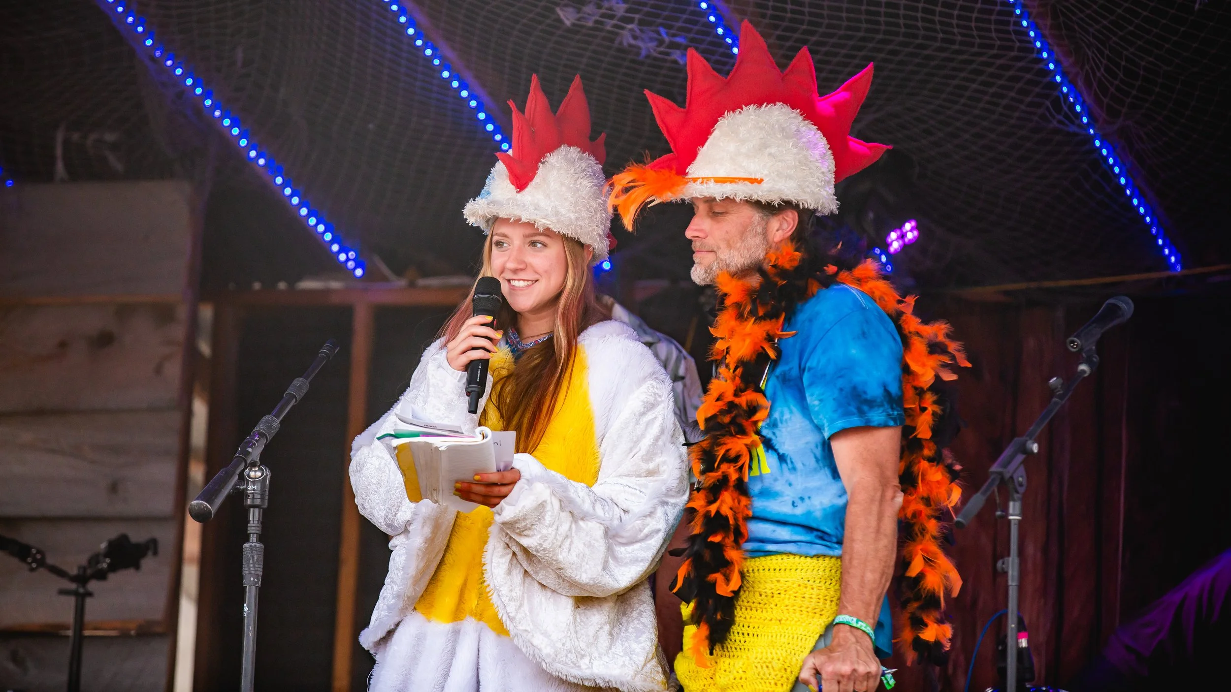 Two people on stage dressed in colorful costumes with large feathered hats, one holding a microphone and notes, the other wearing a feathered boa, in front of a dark background with blue string lights.