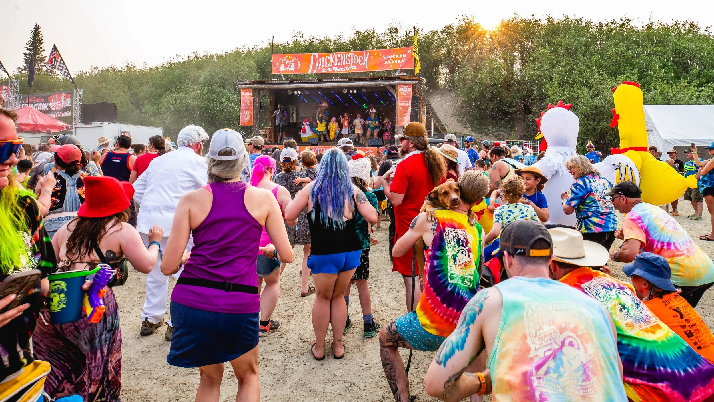Crowd of people at an outdoor music festival with a stage and performers, some dressed in colorful costumes and tie-dye shirts, and inflatable chicken figures, on a sunny day with green trees in the background.