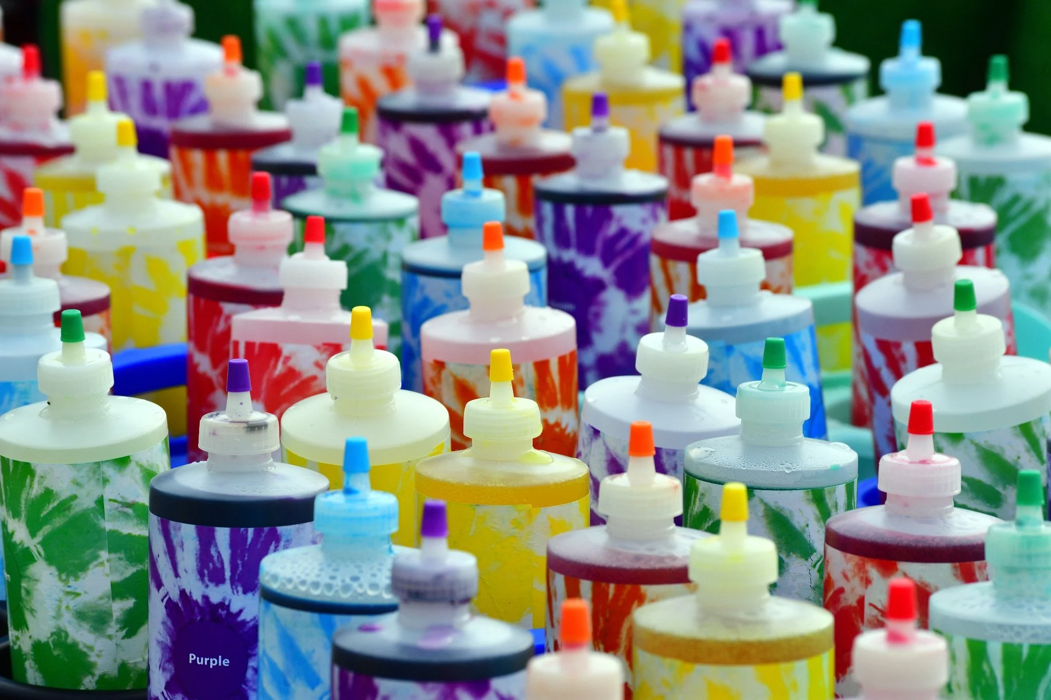 A collection of colorful bottles of paint with caps, arranged on a surface. The bottles have vibrant, tie-dye style designs in various colors including purple, yellow, red, green, blue, black, and white.