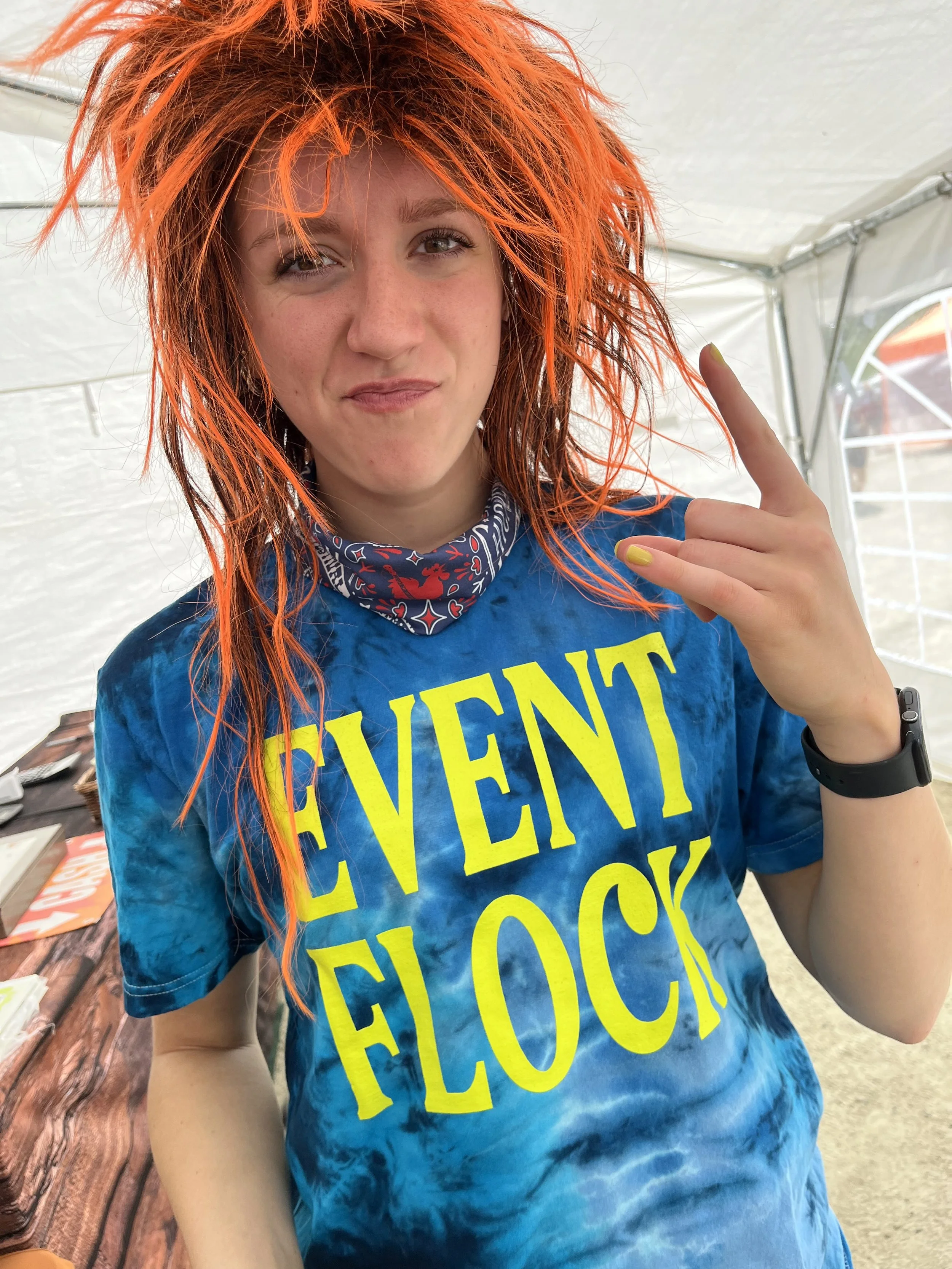 Young woman with bright orange hair, wearing a blue tie-dye shirt with yellow text that says 'EVENT FLOCK'. She is making a rock and roll hand gesture, standing inside a tent with a wooden table in the background.
