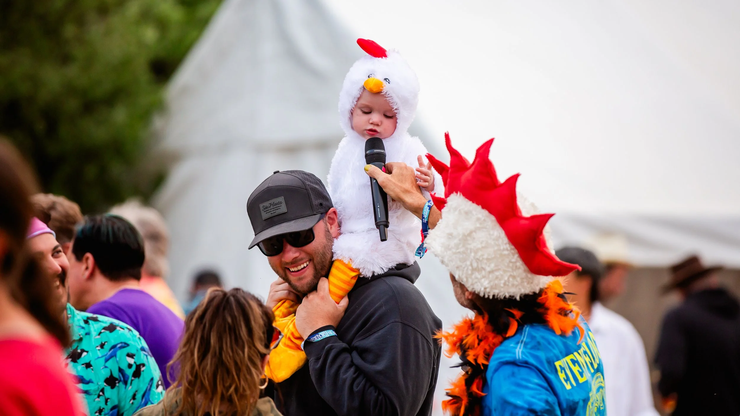Children dressed in costumes at a festive event, with one child in a chicken costume holding a microphone, seated on a man's shoulders, and another person in a dragon costume nearby, amidst a crowd.