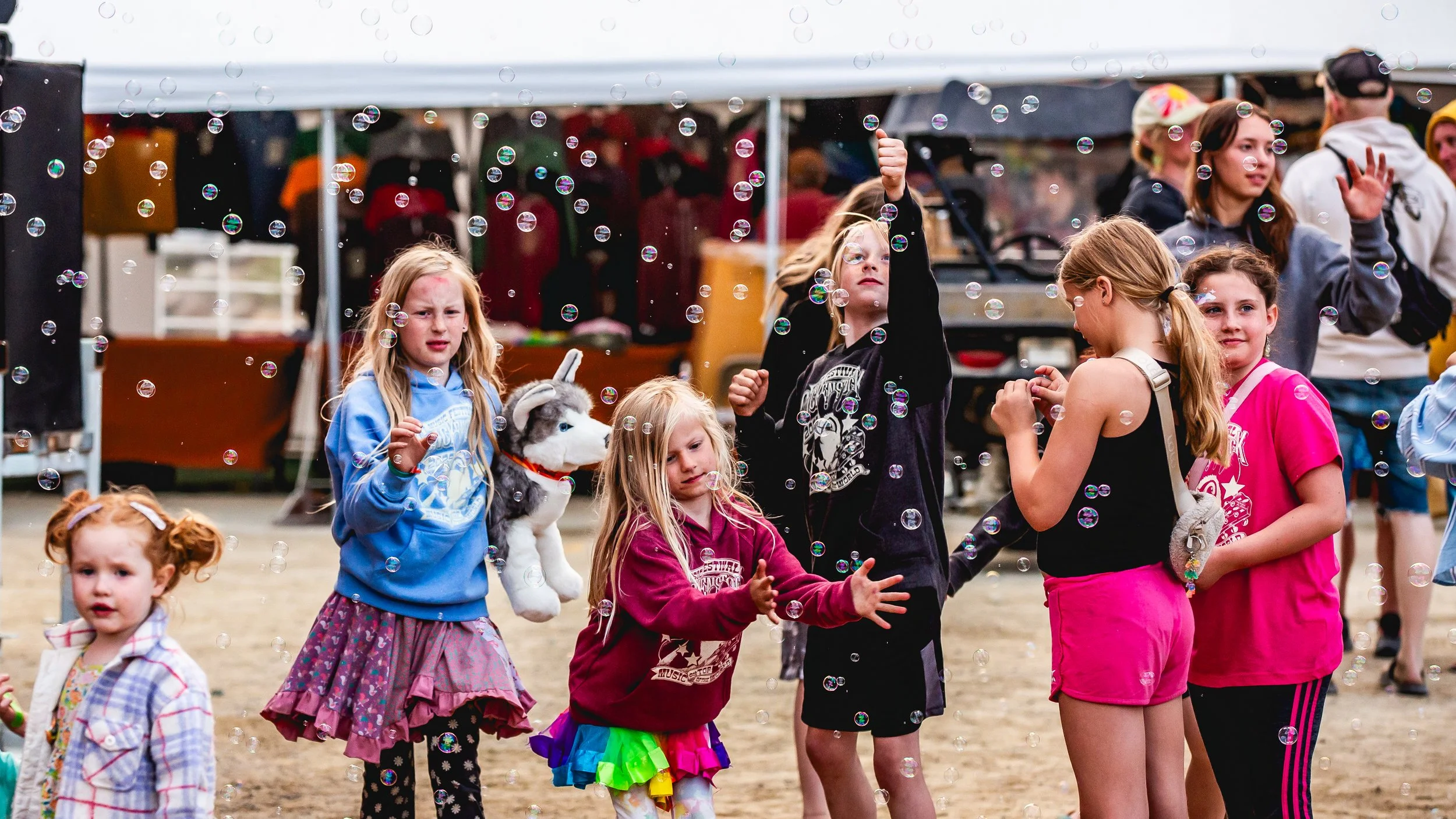 Group of children playing with bubbles at an outdoor fair or festival, some running and reaching for bubbles, with booths and people in the background.