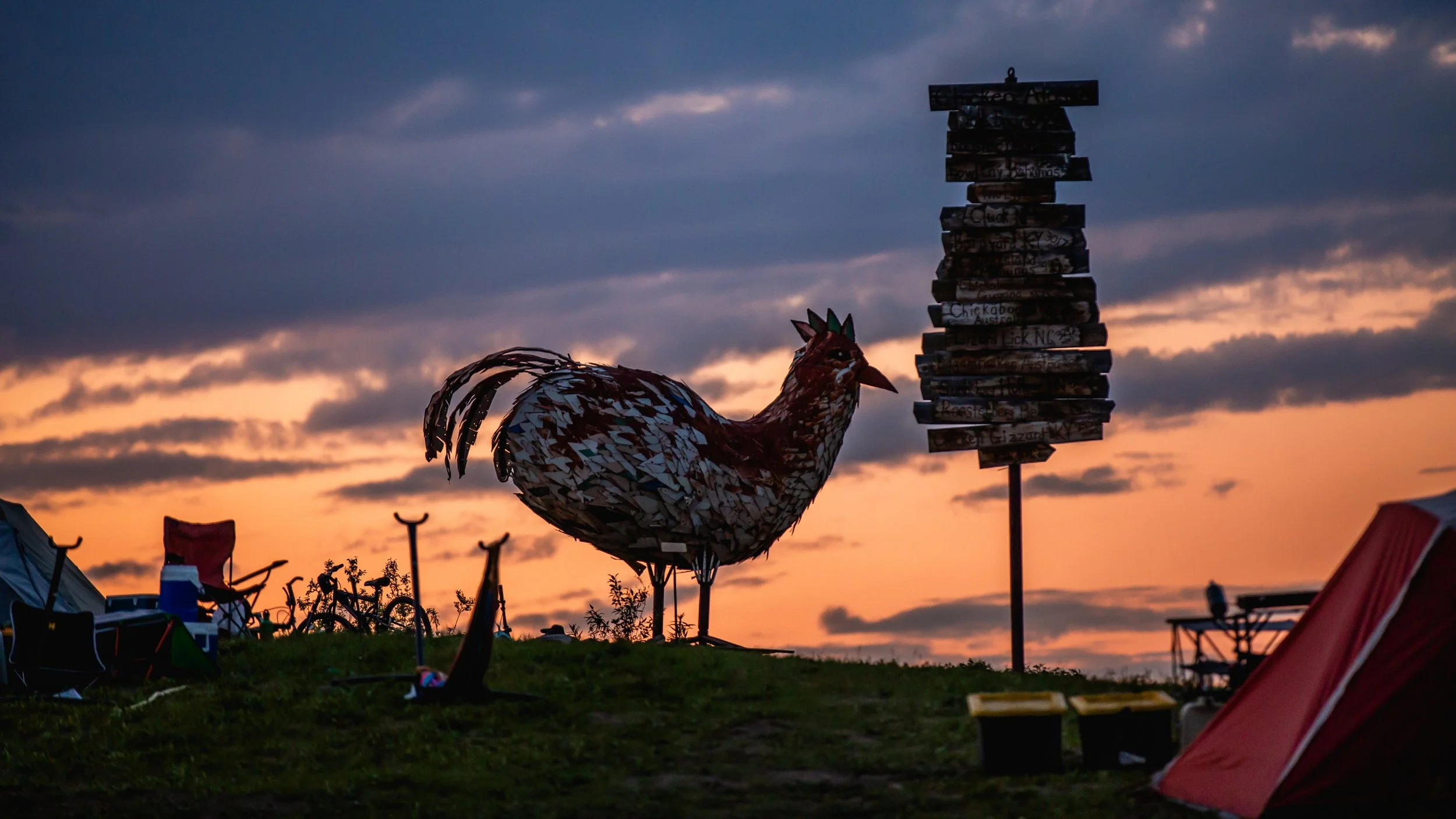 A large rooster sculpture made of metallic pieces and a sign made of stacked wooden planks on a grassy hill at sunset, with tents and camping chairs in the background.