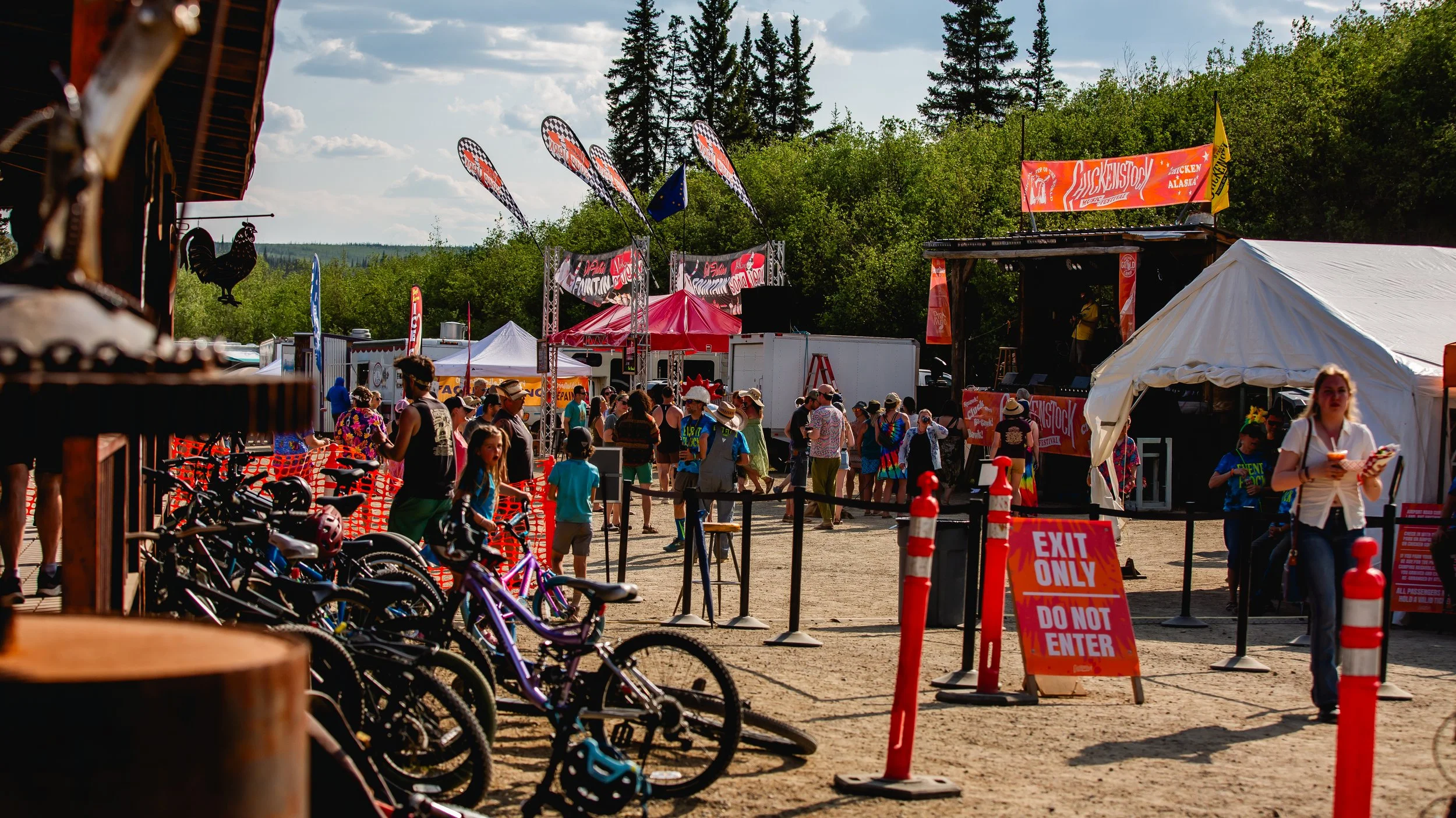 Crowd of people at an outdoor festival with bicycles, tents, and a stage. There are trees and a partly cloudy sky in the background.
