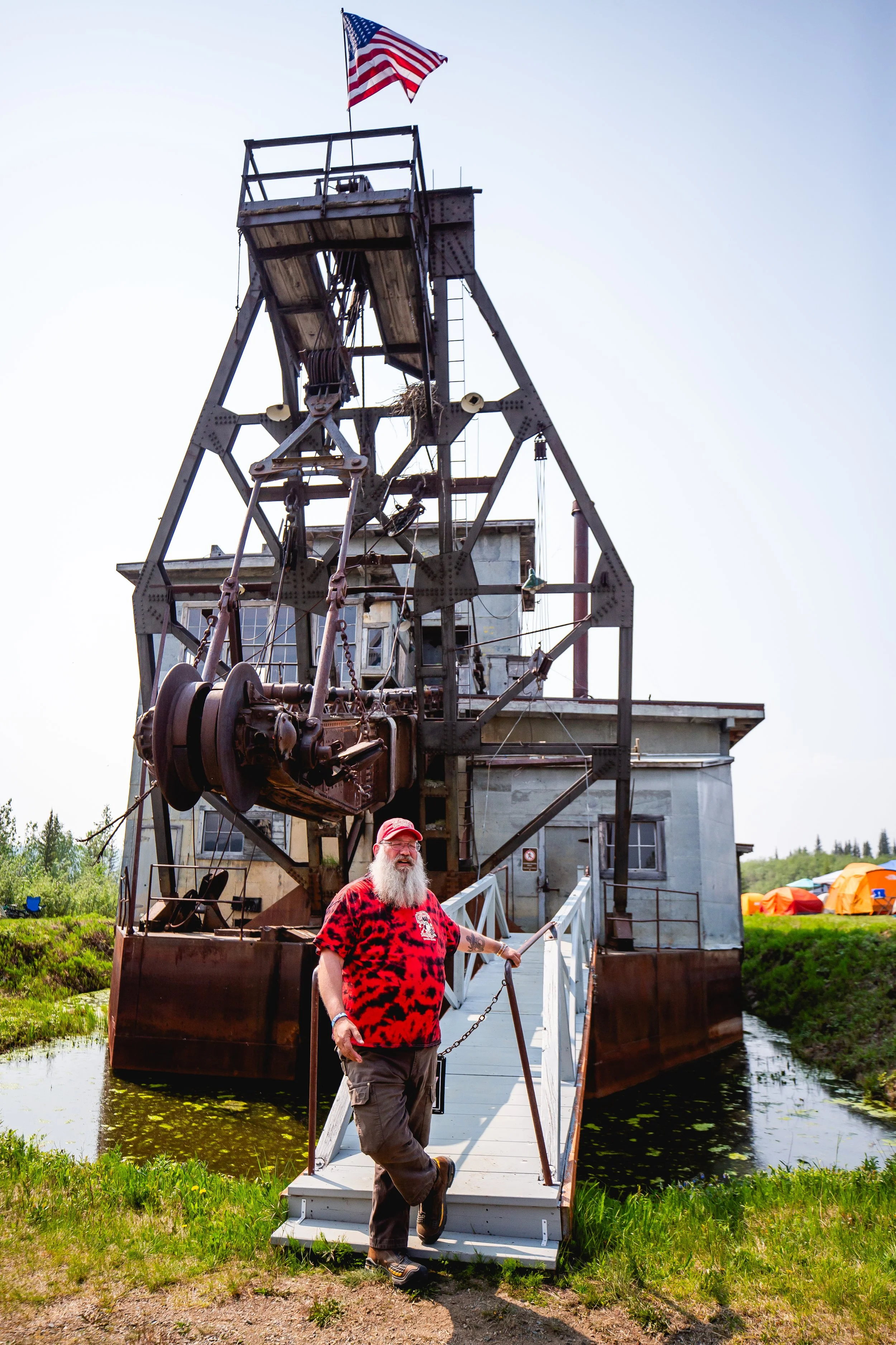 A man with a long white beard, wearing a red and black leopard print t-shirt and a red cap, standing on a small dock in front of a large, old industrial crane structure, with an American flag on top.