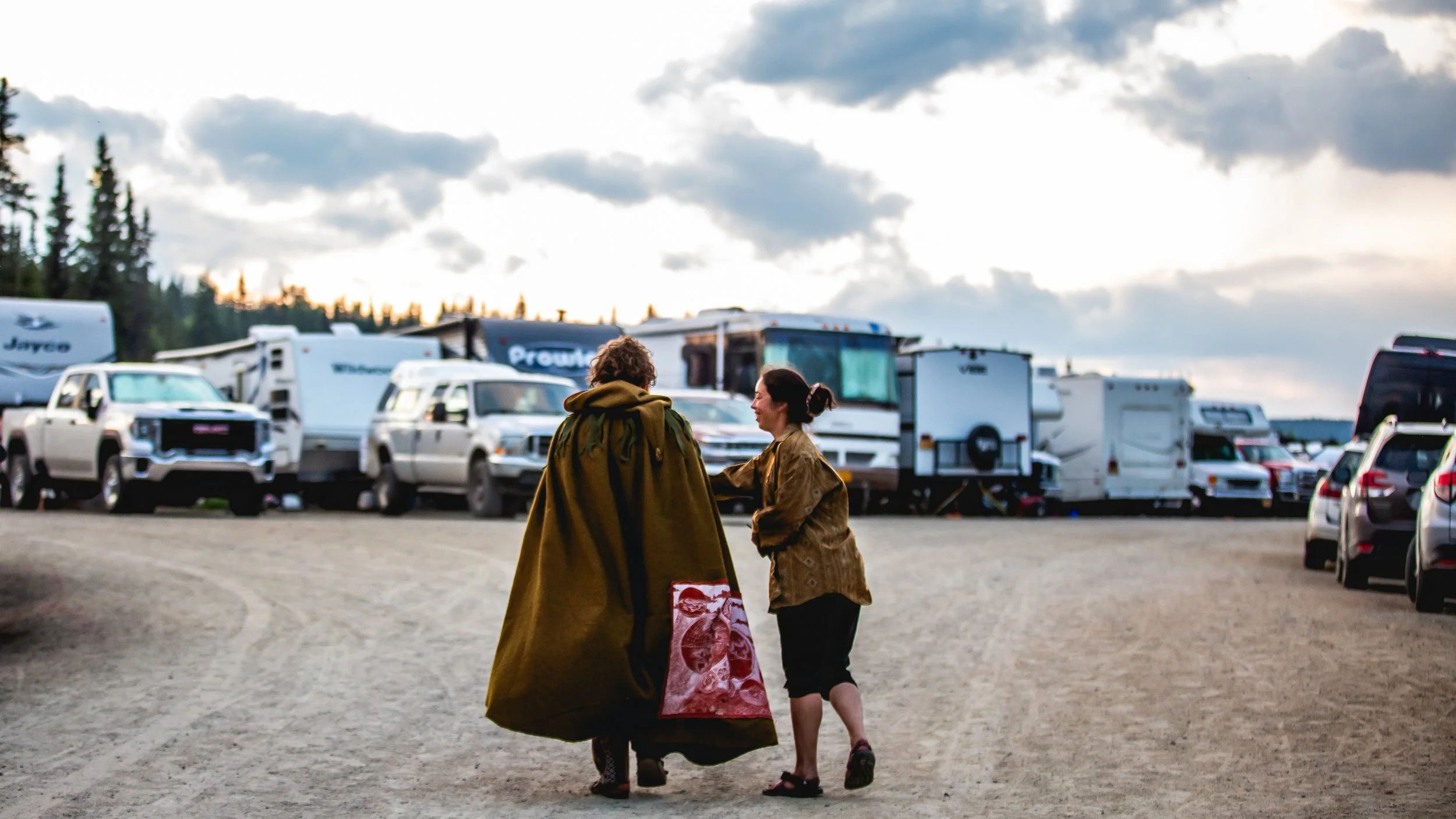 Two people walking together in a parking lot filled with RVs and cars, with a cloudy sky above.