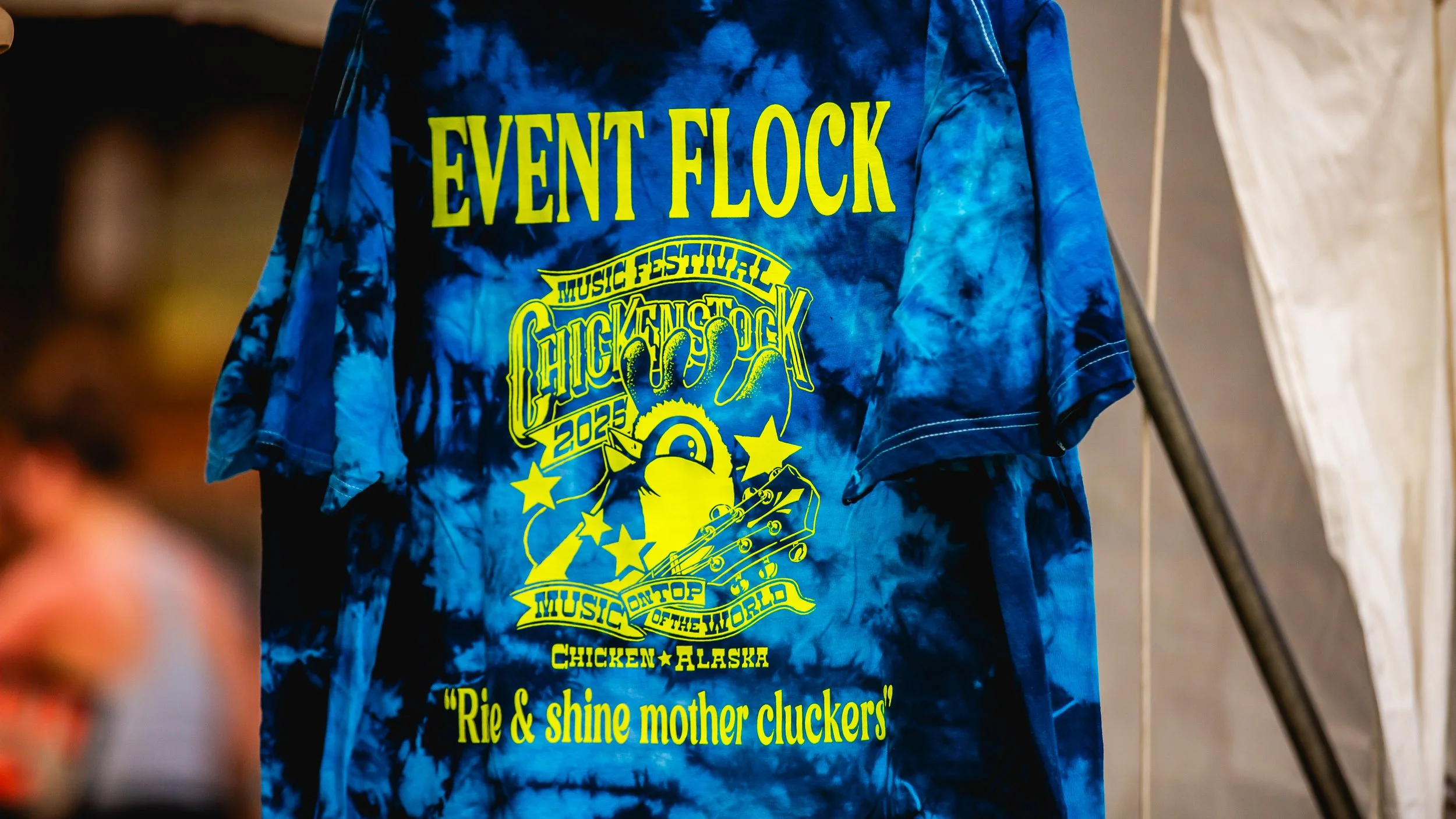 Blue tie-dye T-shirt with yellow text and graphics promoting the 2023 Chuck Fest music festival in Chicken, Alaska, including a guitar, stars, and a chick illustration.