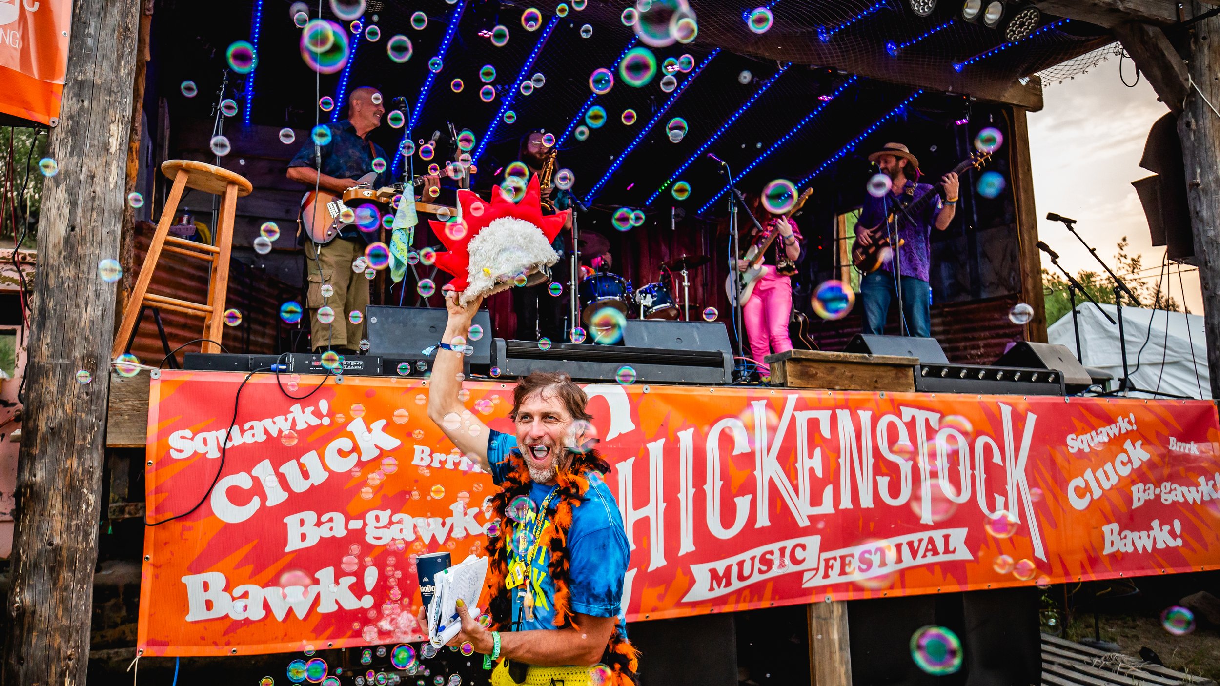 A lively outdoor music festival with a stage featuring a band playing guitars and drums, decorated with neon lights and bubbles. A man in a blue shirt with a lei and a feather boa is smiling and holding a costume headpiece shaped like a chicken, in front of a large orange banner that reads 'Hickenstock Music Festival.' The sky suggests it is during evening or sunset.