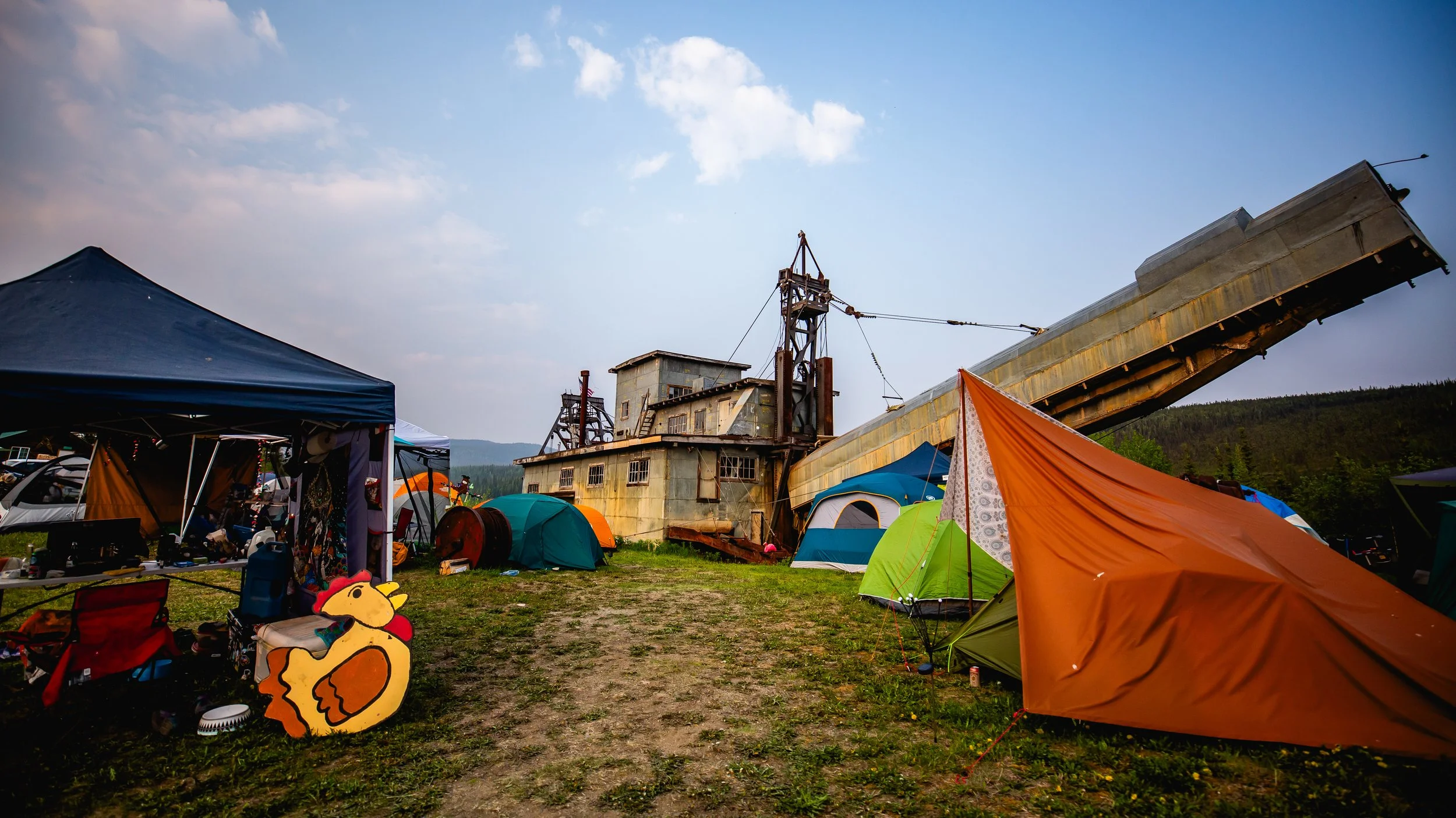 A campsite with tents and an abandoned mining or industrial structure in a rural area with mountains in the background.