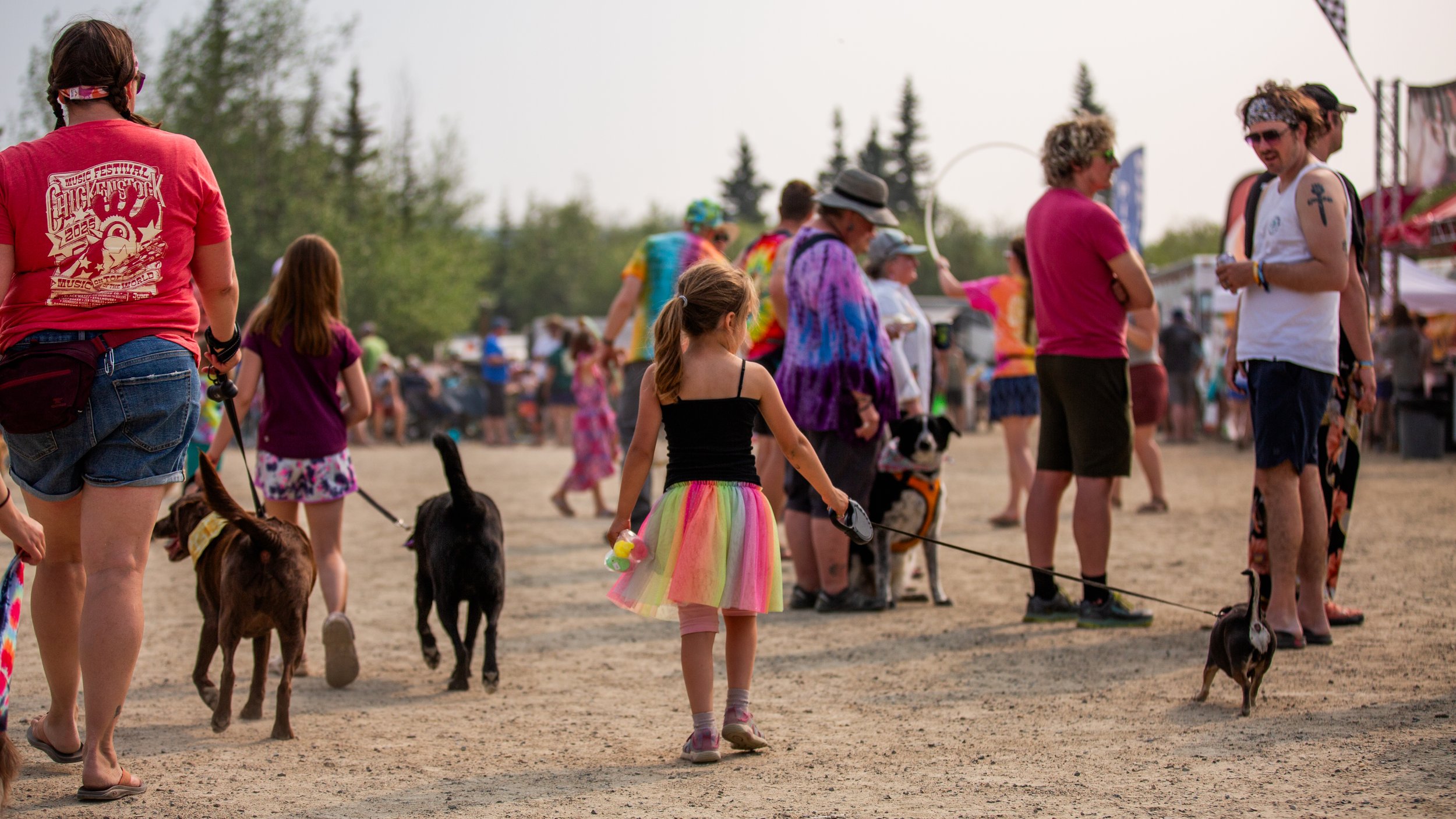 People walking and socializing at an outdoor festival, some with dogs, little kids, and colorful attire on a dusty ground with trees and vendor booths in the background.