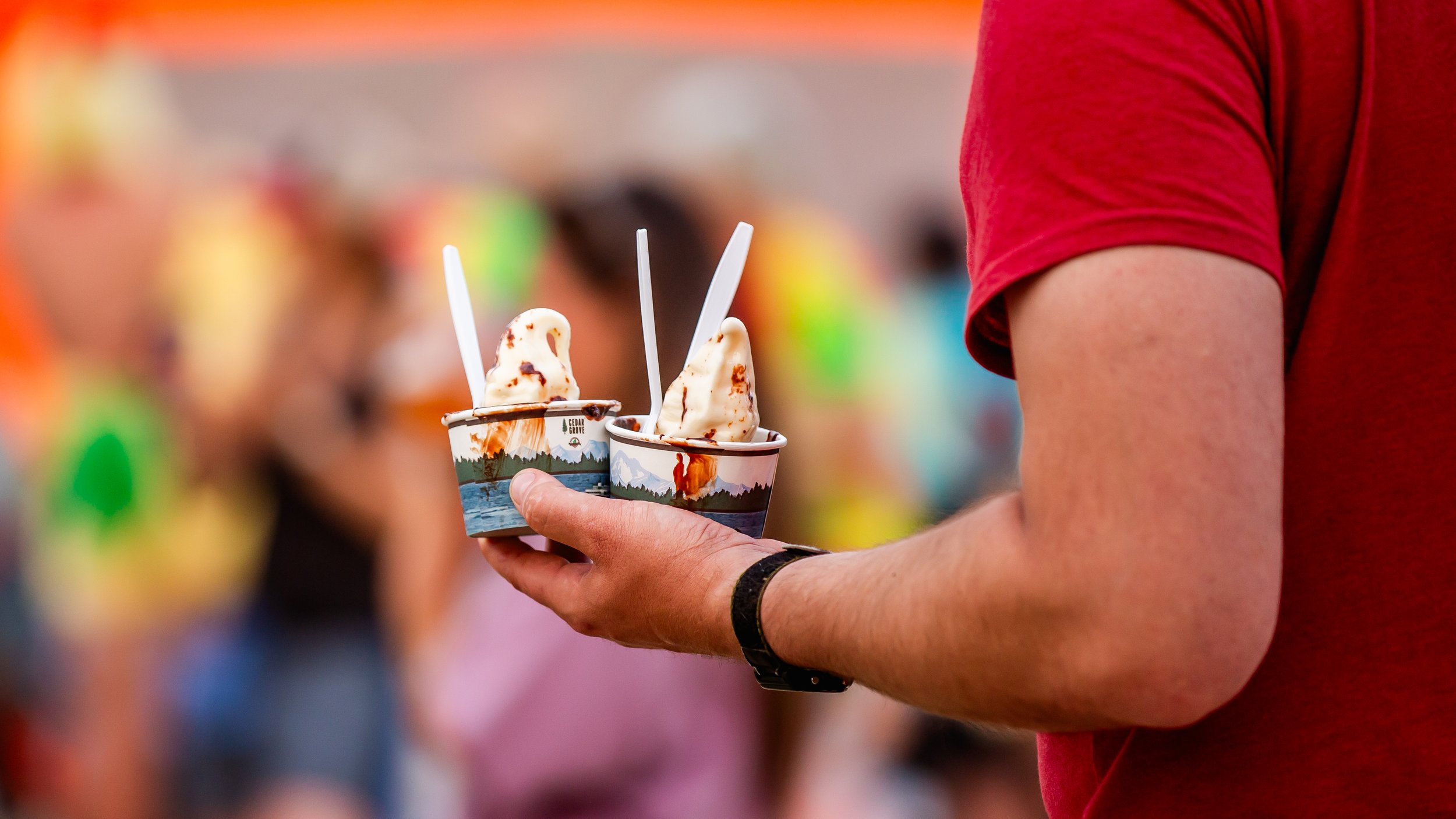 Person holding two cups of ice cream at what appears to be a busy outdoor event.