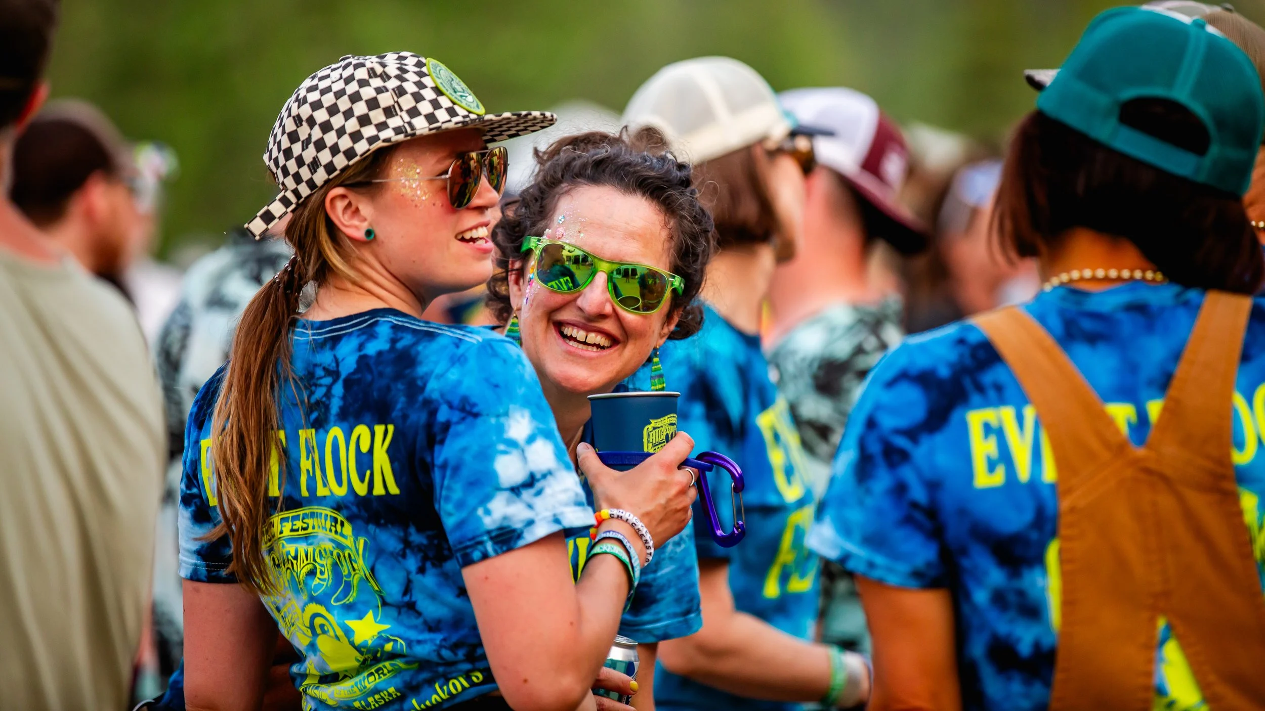 A group of people wearing blue tie-dye shirts with yellow lettering, attending an outdoor festival. Two women in the foreground are smiling and wearing sunglasses, with one holding a drink cup.