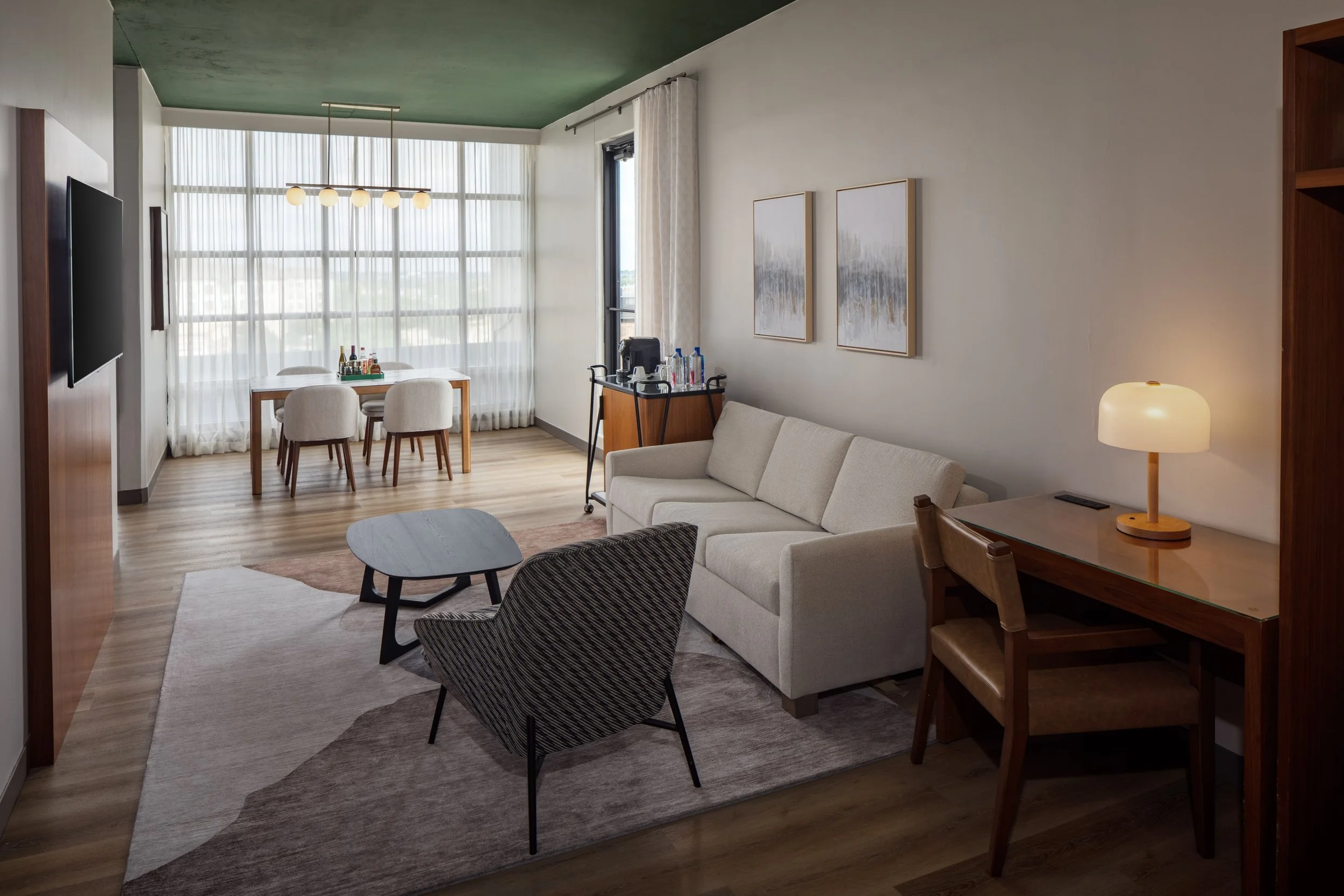 Living area of the 1 King Bed One Bedroom Suite with Balcony at The Clara Hotel in Plano, Texas, featuring a workspace, sofa, dining table, minibar, HDTV, and balcony access.