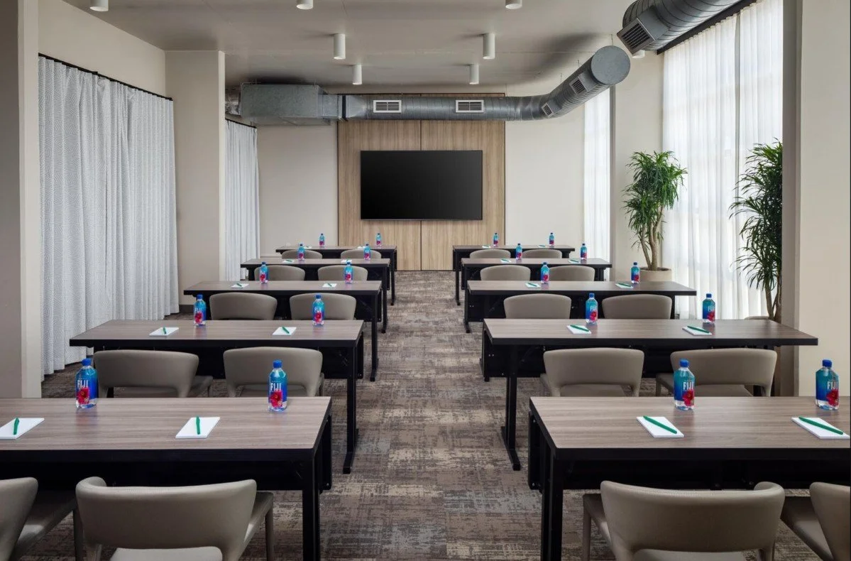 Legacy Meeting Space at The Clara Hotel in Plano, Texas, with a classroom-style setup, a 100-inch smart TV, and tables set with Fiji water, notepads, and pens.