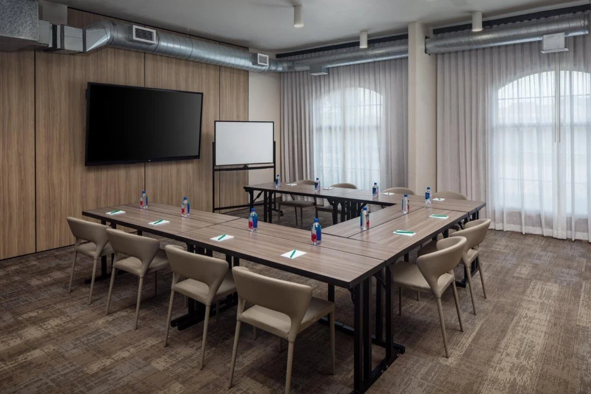 Plano Meeting Space at The Clara Hotel in Plano, Texas, with a U-shaped setup, a smart flat-screen TV, a whiteboard, and tables set with Fiji water, notepads, and pens.