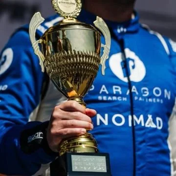 Person holding a large gold trophy in front of a blue jacket with patches.