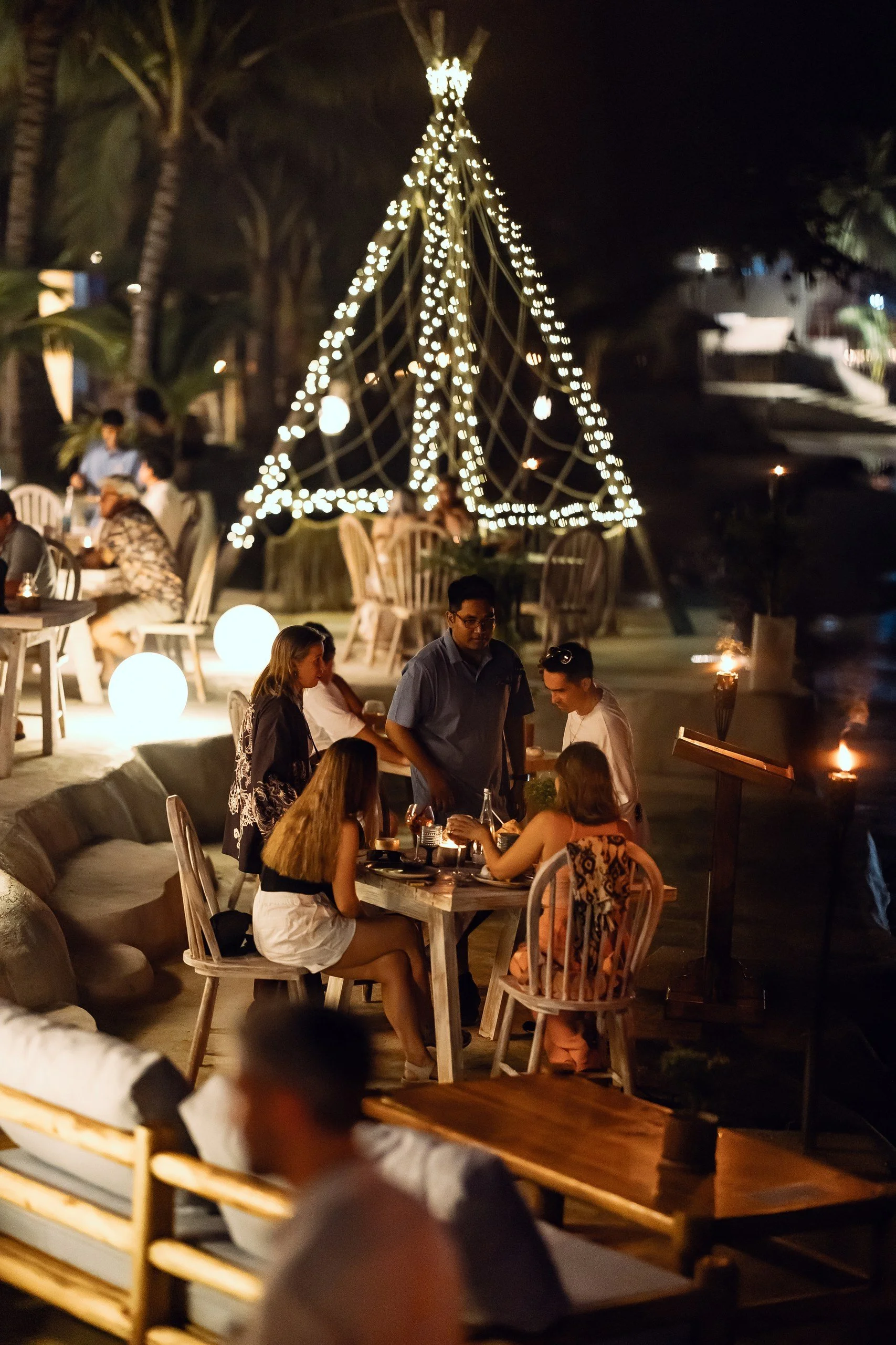 People dining outdoors at night near a decorated with string lights and a large illuminated cone-shaped structure.